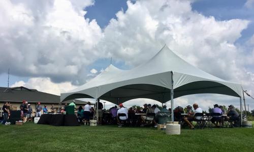 Large white tent with open sides shields golfers, seated at tables, from the sun at the Olathe Golf Tournament