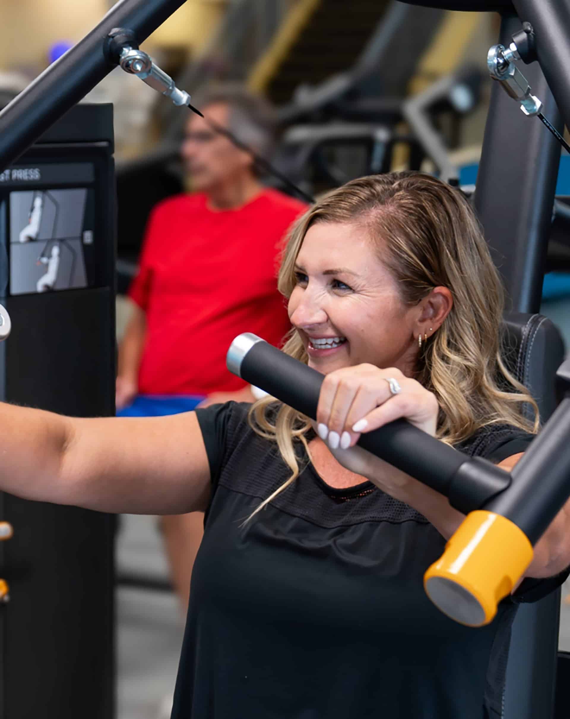 Smiling woman using a chest-press machine in the YMCA fitness center with another member nearby