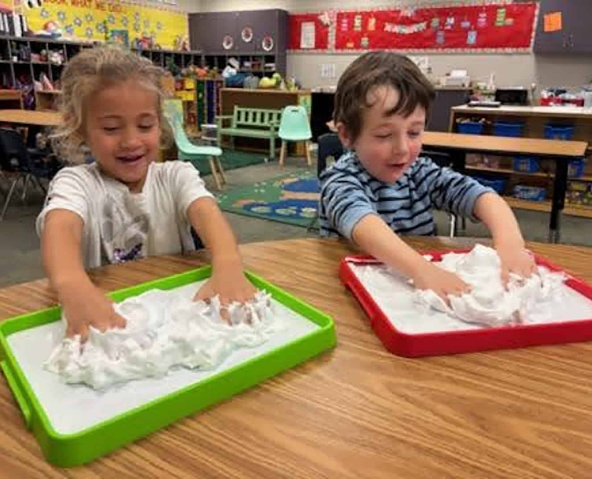 Two young children exploring shaving-cream sensory trays at a YMCA preschool classroom table