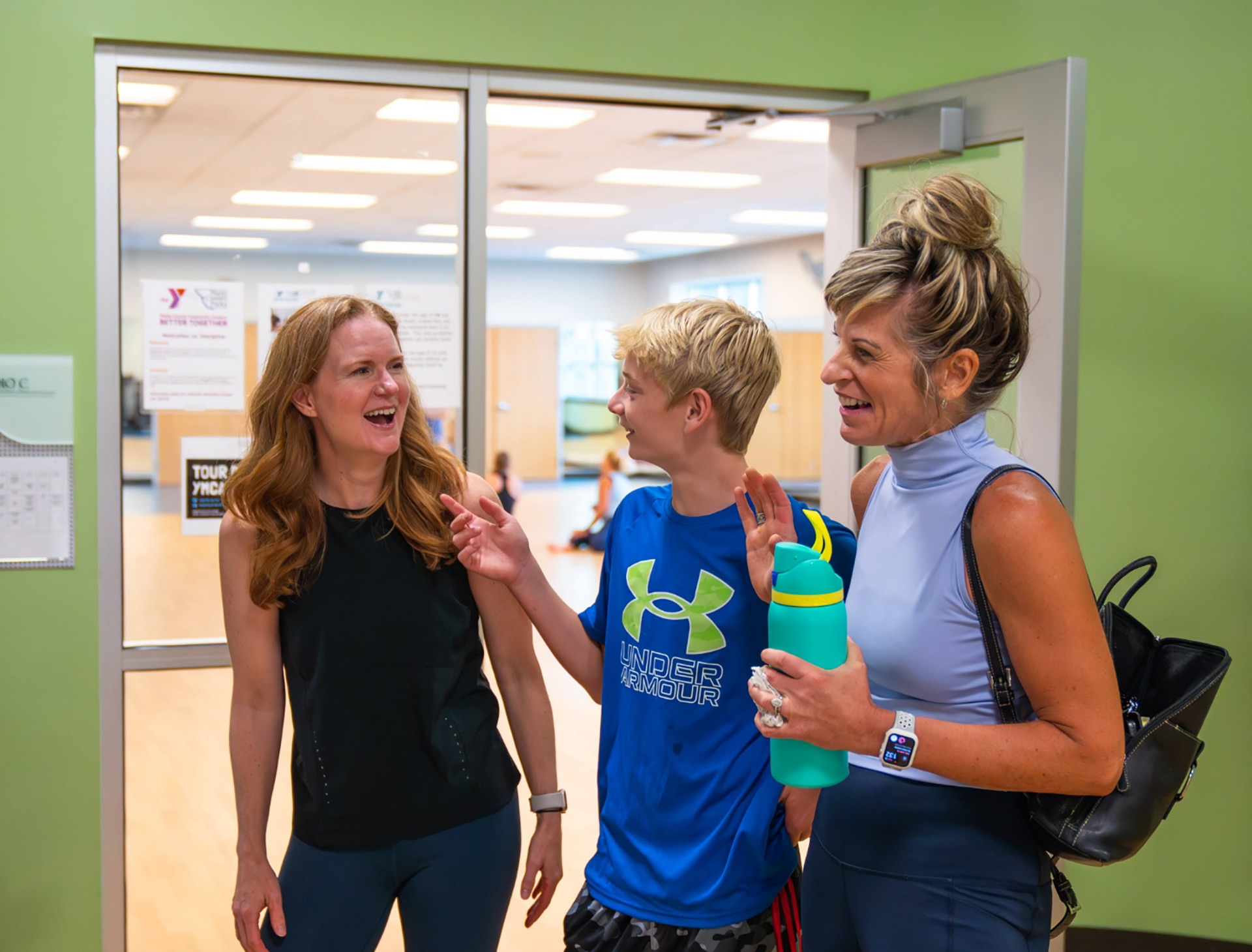 Woman, teen boy, and woman laughing together at the entrance of a YMCA fitness studio