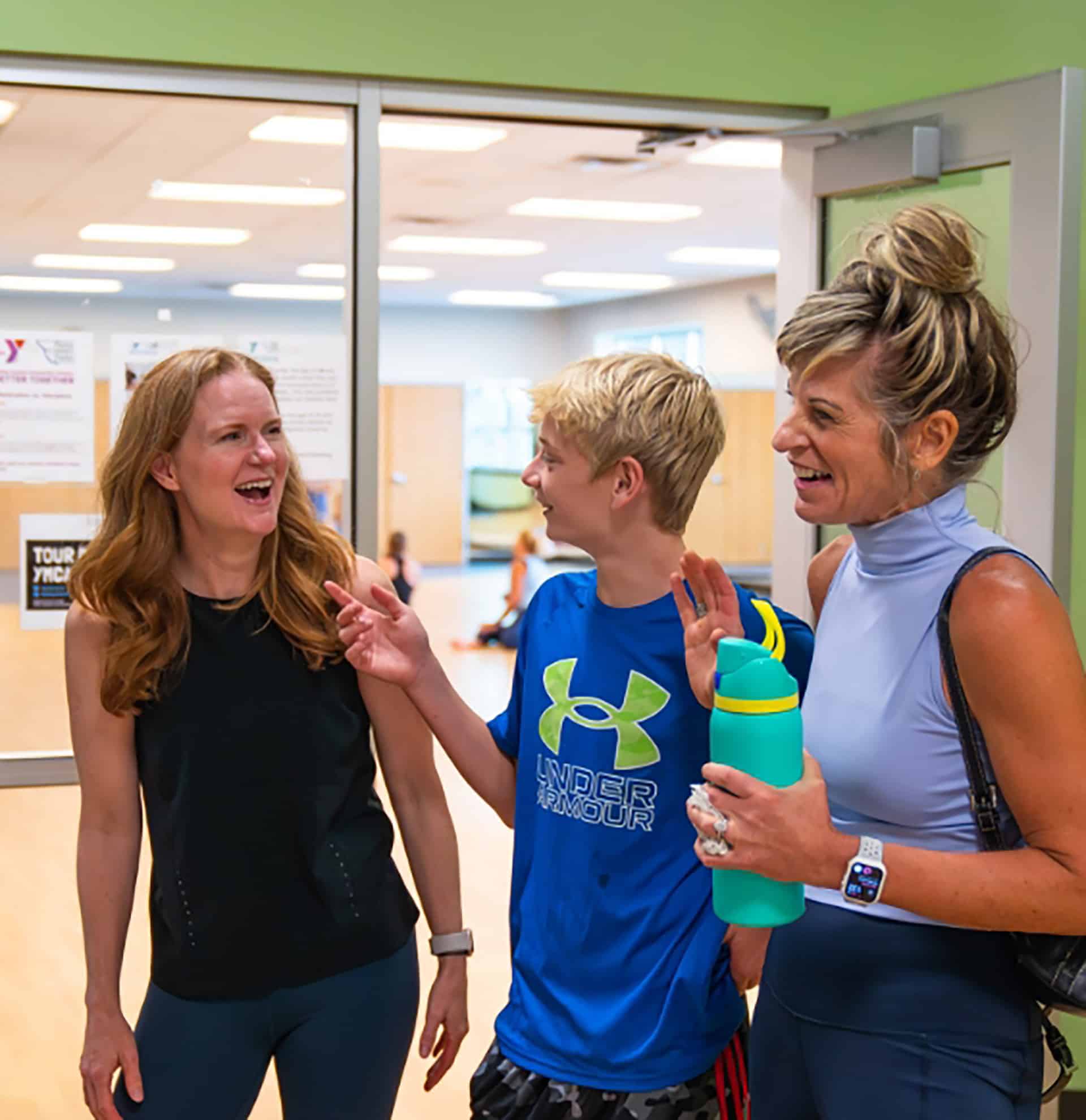 Woman, teen boy, and woman laughing together inside a bright YMCA wellness center