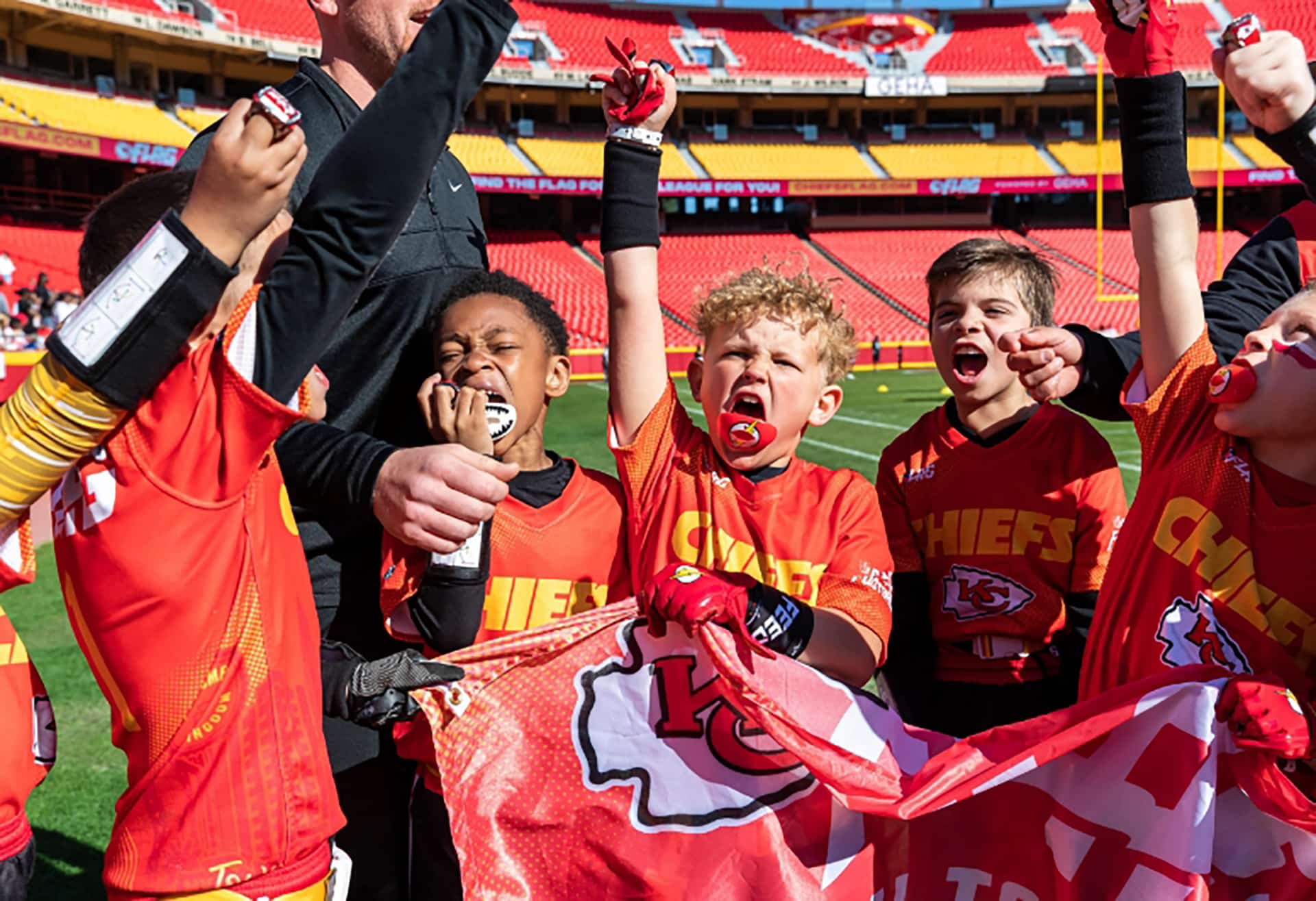 Kids in Chiefs flag football jerseys cheering with a Kansas City Chiefs flag at the stadium
