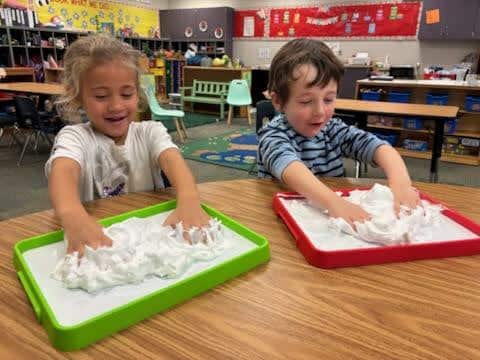 Two preschoolers laughing while playing with shaving cream on trays in a classroom