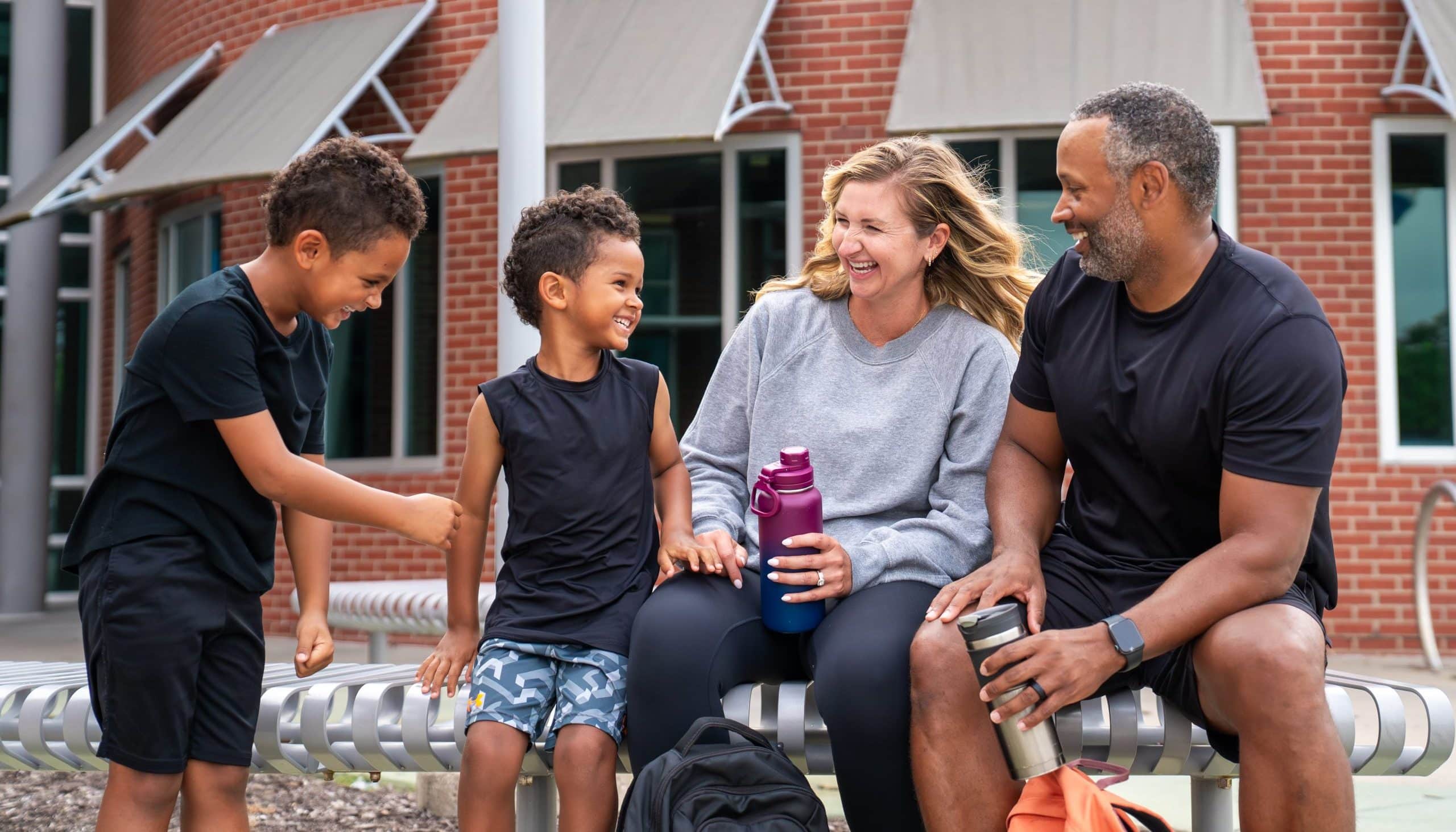 Family of four laughing together on an outdoor bench outside a brick YMCA building