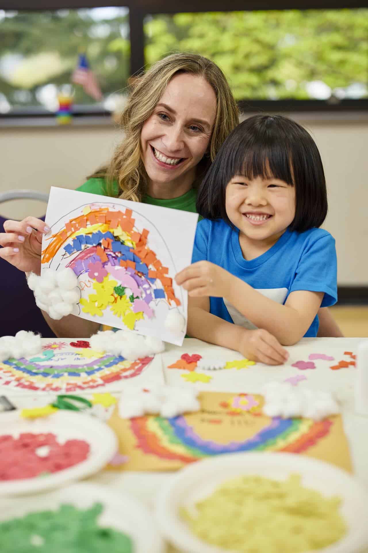 Smiling woman and young child holding up a colorful rainbow craft at a table