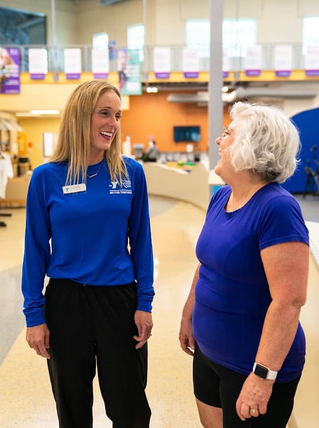 YMCA staff member in blue shirt laughing with an older woman member on the wellness floor