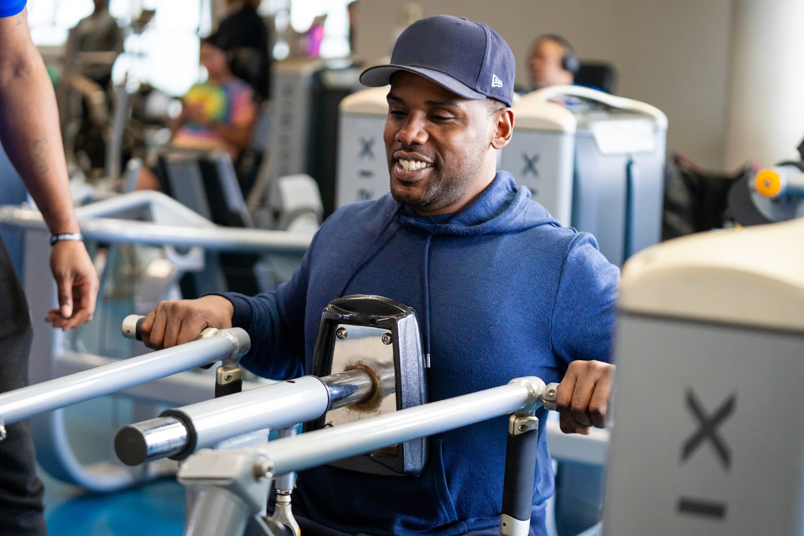 Smiling man in a navy hoodie and cap using a chest press machine on a busy gym floor