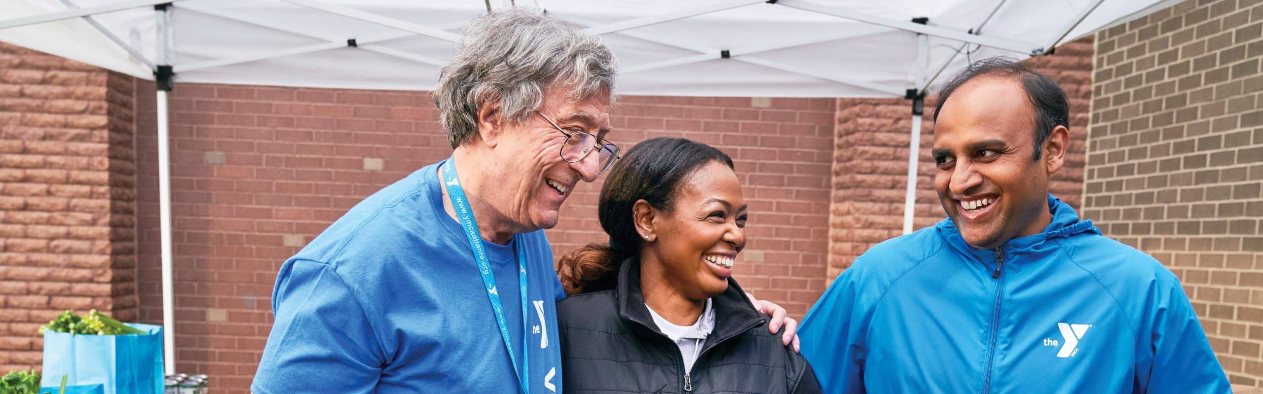 Three YMCA volunteers in blue jackets laughing together under a tent at an outreach event