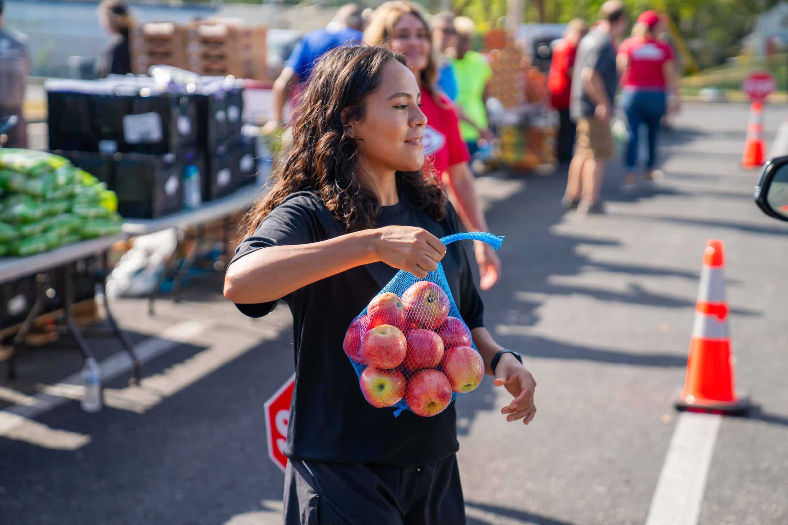 Smiling volunteer carrying a mesh bag of apples at an outdoor community food distribution event