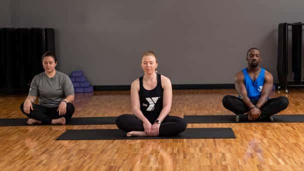 Three adults meditate cross-legged on yoga mats in a calm studio