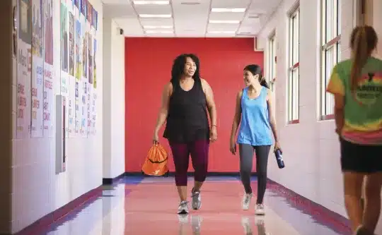 Two women in workout clothes walking and chatting down a bright YMCA hallway with a red accent wall