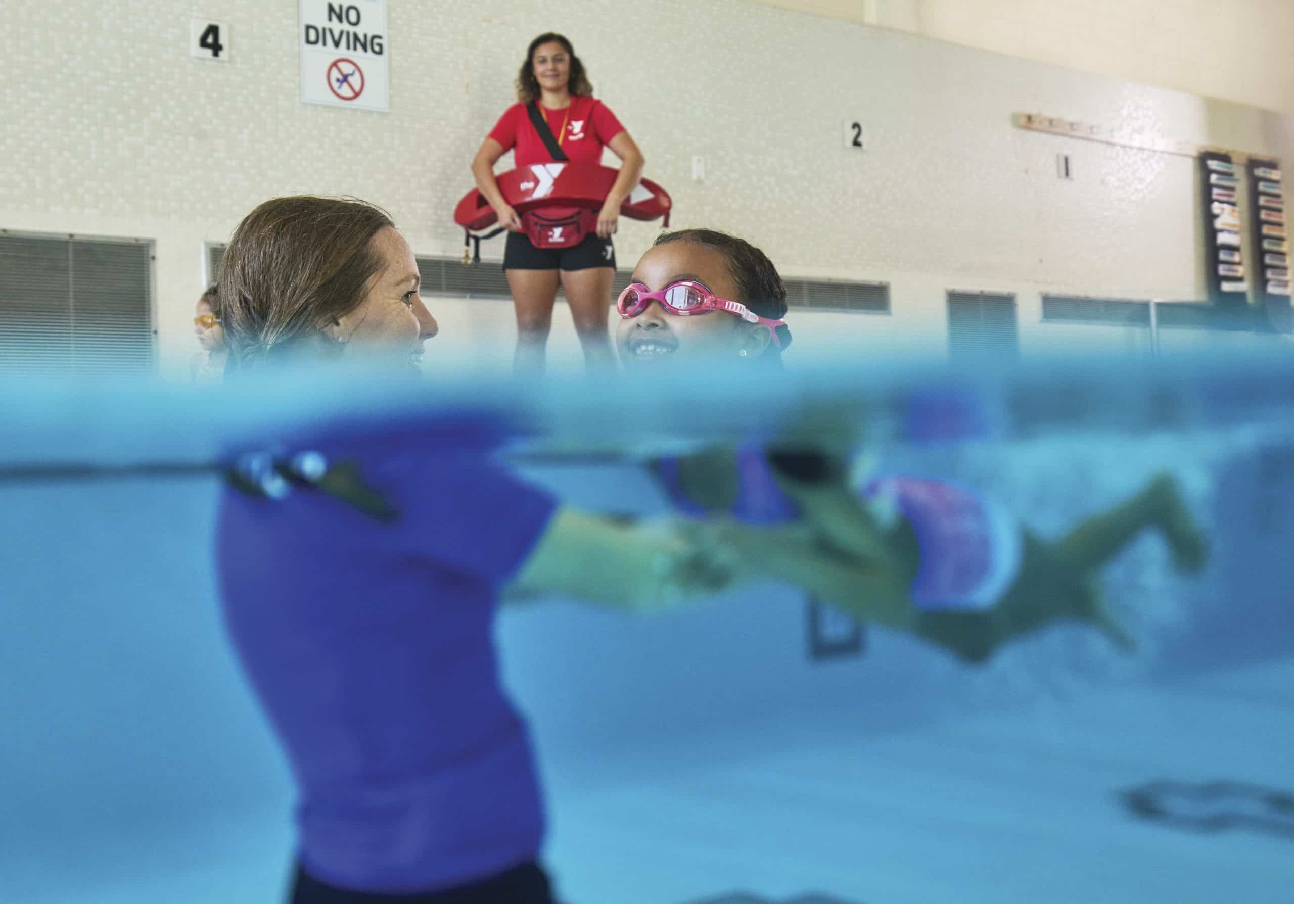Instructor guides a young child in pink goggles swimming in an indoor pool