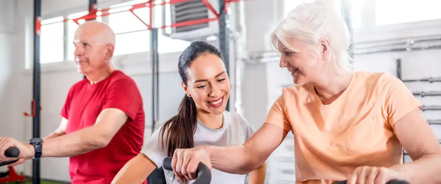 A trainer smiles with two older adults riding stationary bikes in a fitness studio