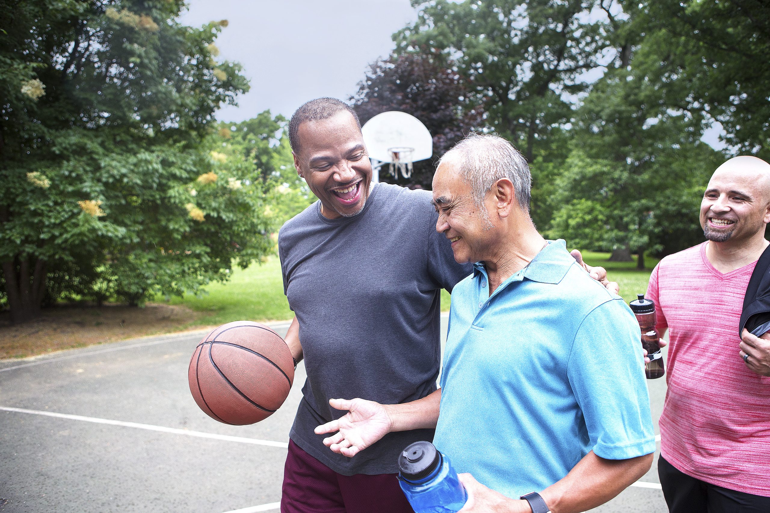 Three men laugh together on an outdoor basketball court, one holding a basketball with an arm around a friend