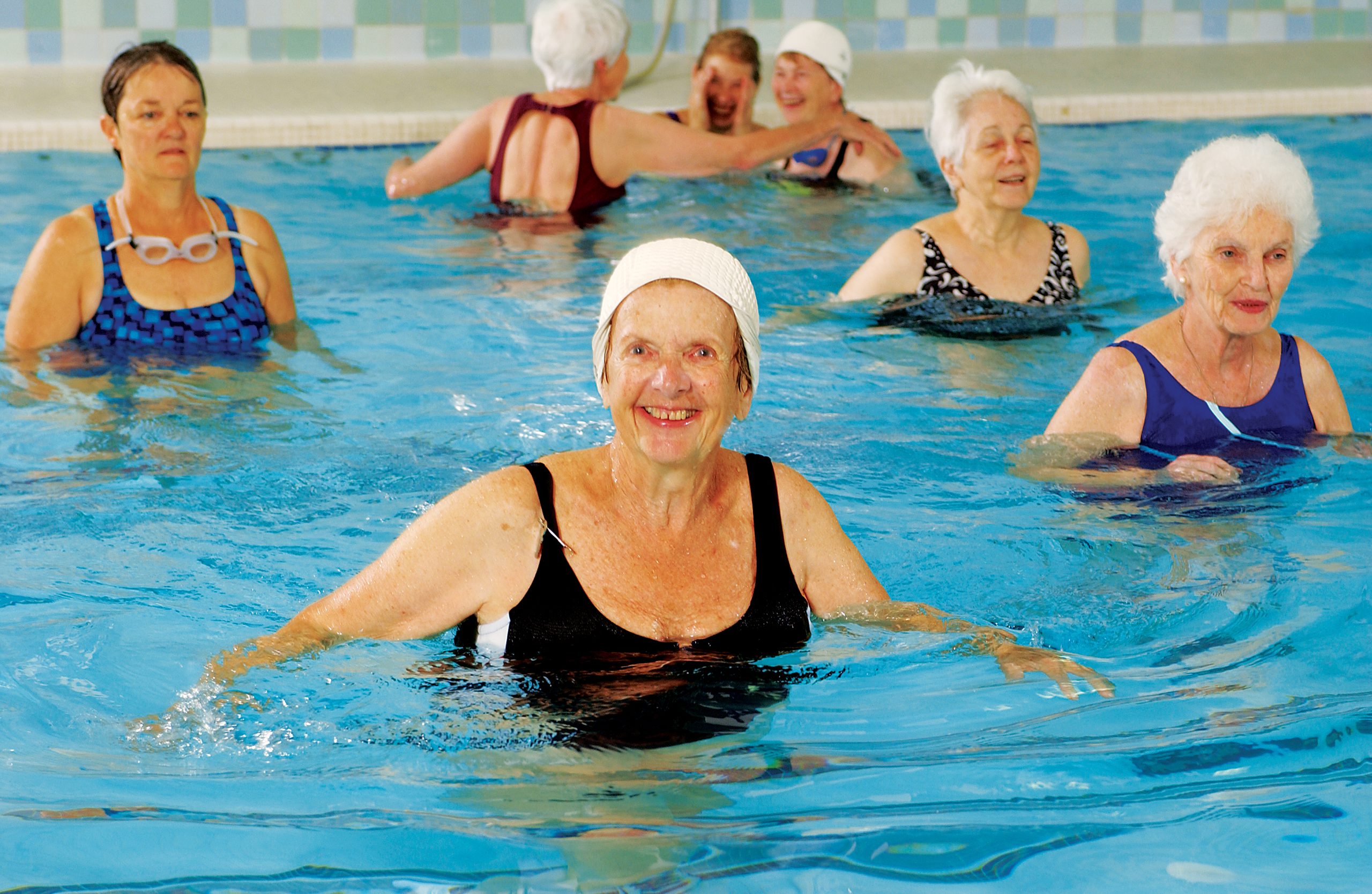 Smiling older adults in swim caps stand chest deep in an indoor pool during a water exercise class