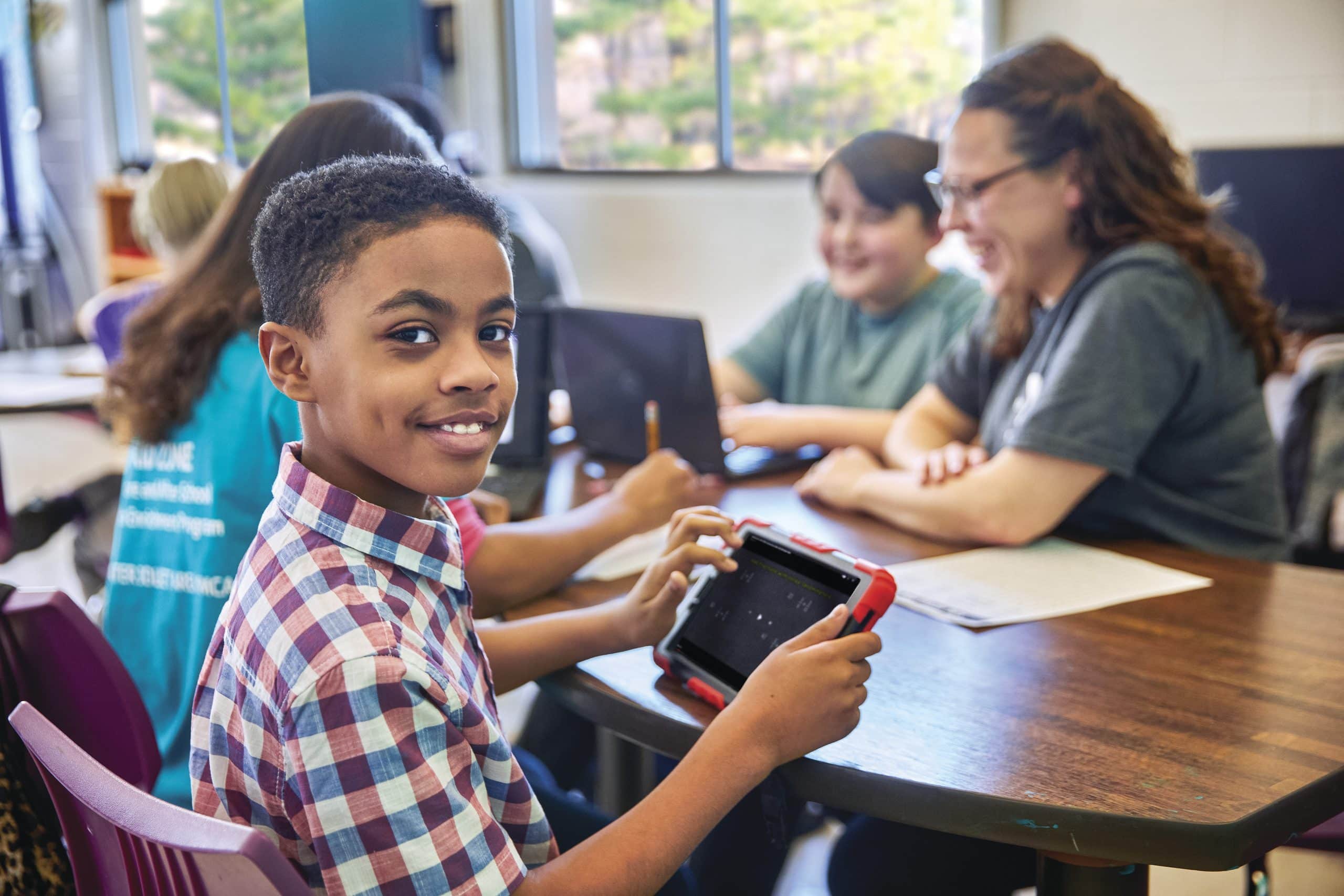 Boy smiling over his shoulder while using a tablet at a table with staff and other students