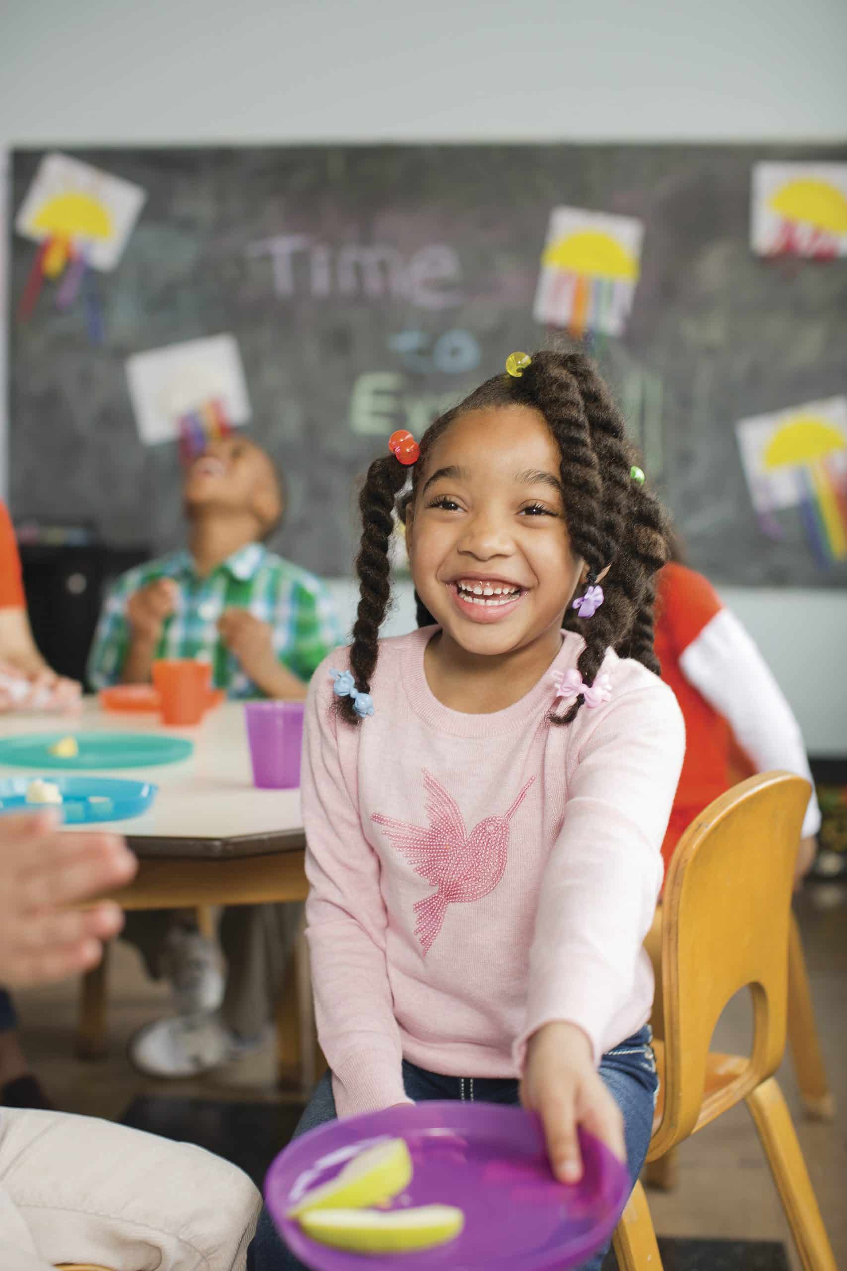 A grinning child with beaded braids holds out a purple plate of apple slices at a classroom snack table