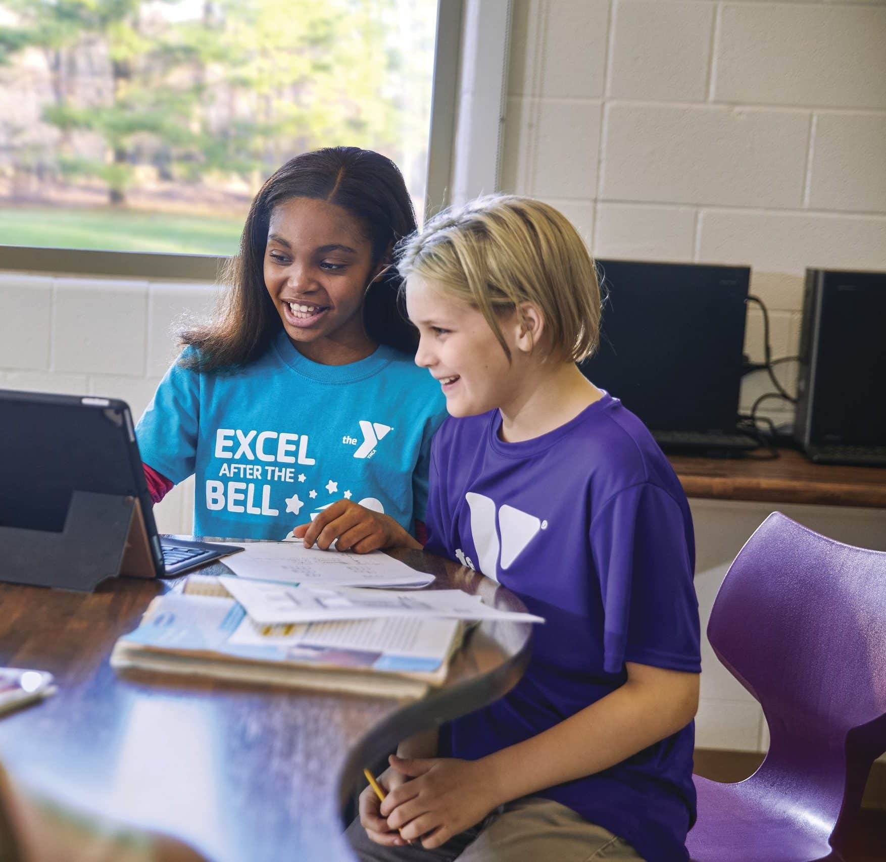 Two students smile at a tablet propped on a table while working through homework together
