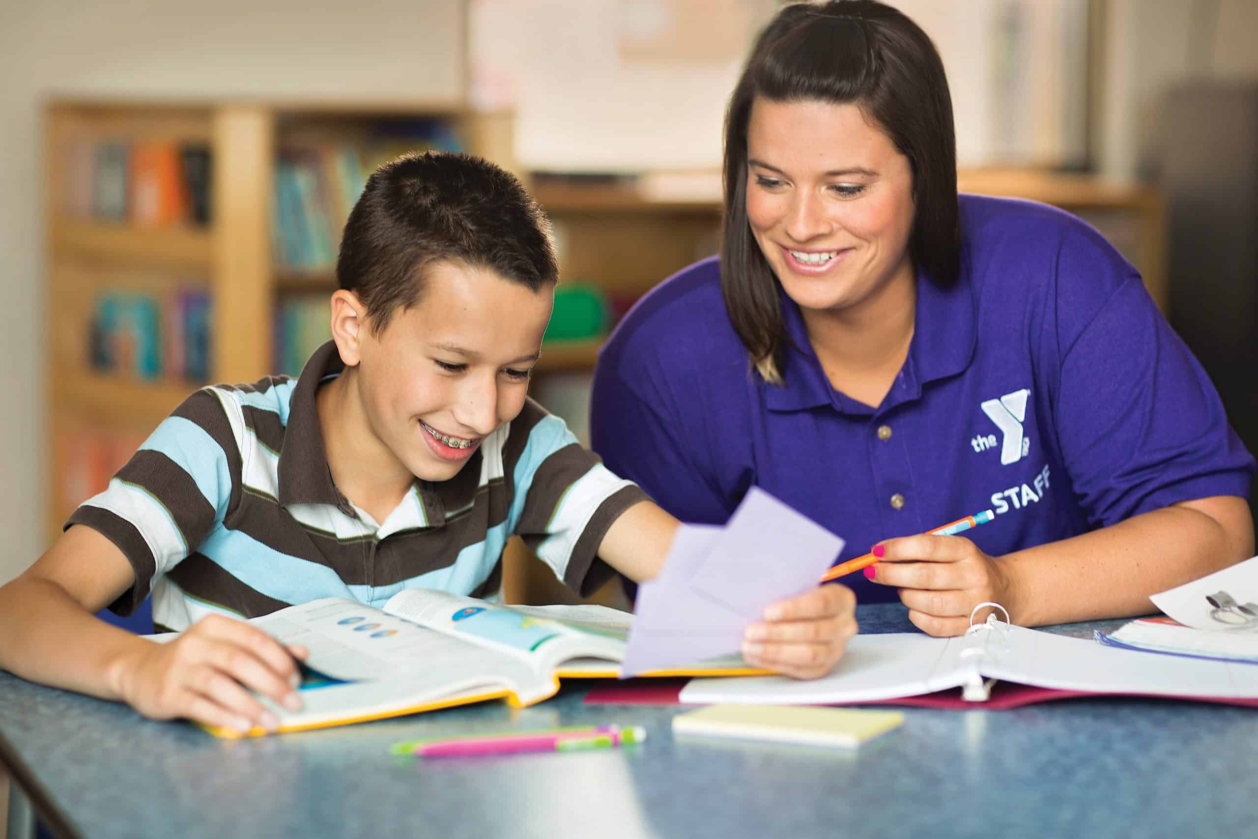 YMCA staff member in a purple polo helps a boy with an open workbook at a classroom table