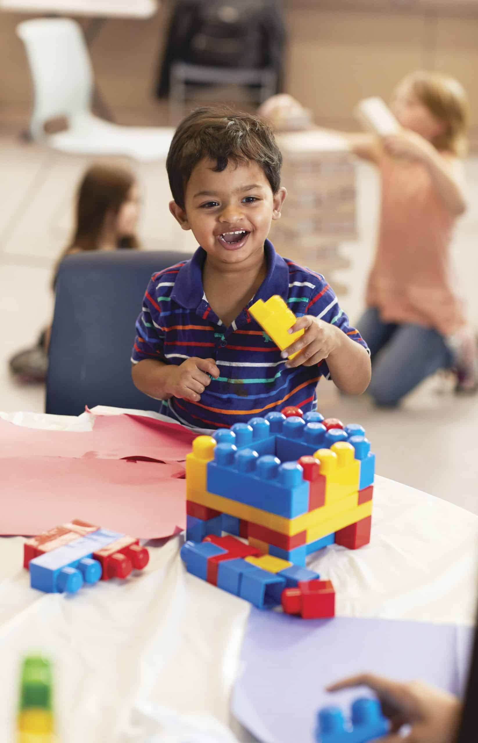 Grinning toddler building a tower with large interlocking plastic blocks at a paper covered table