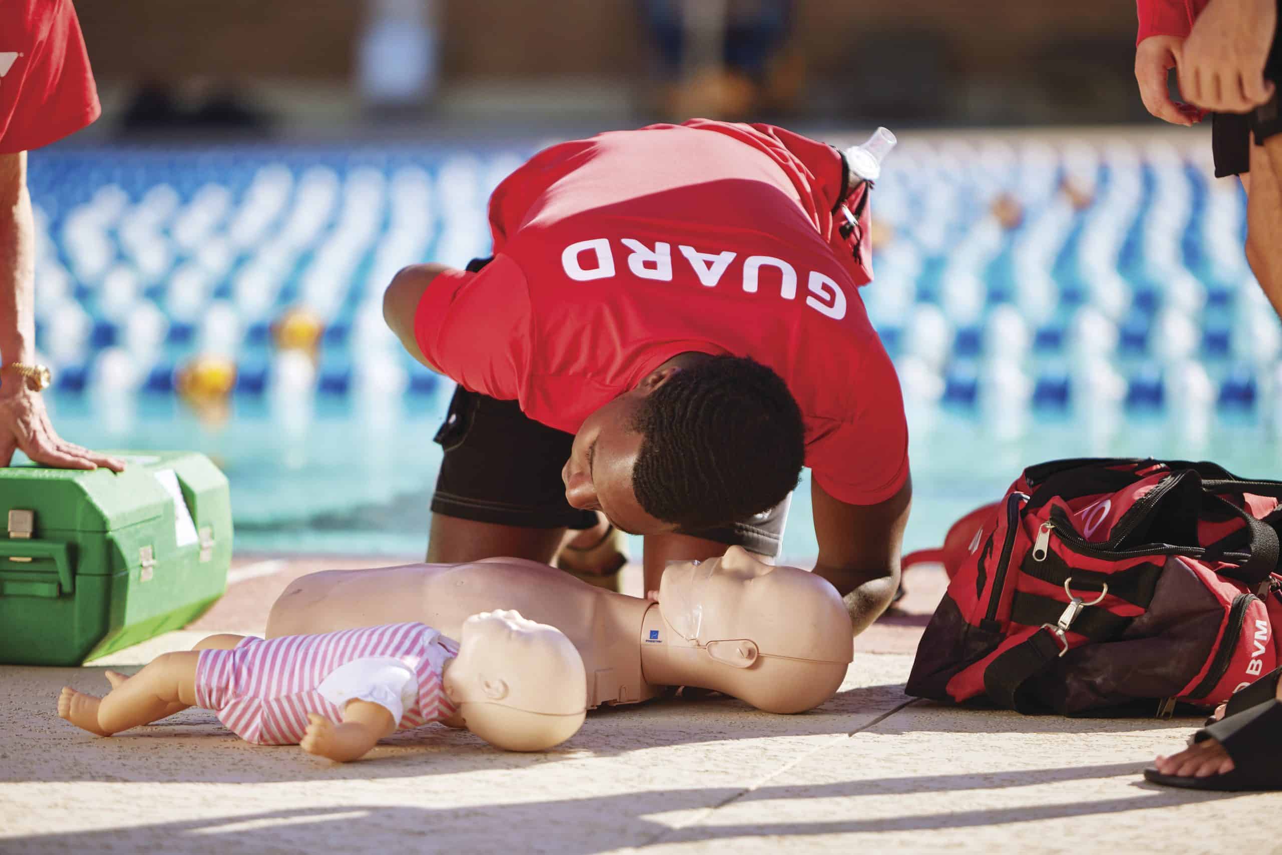 A lifeguard in a red guard shirt practices rescue breathing on adult and infant CPR manikins poolside
