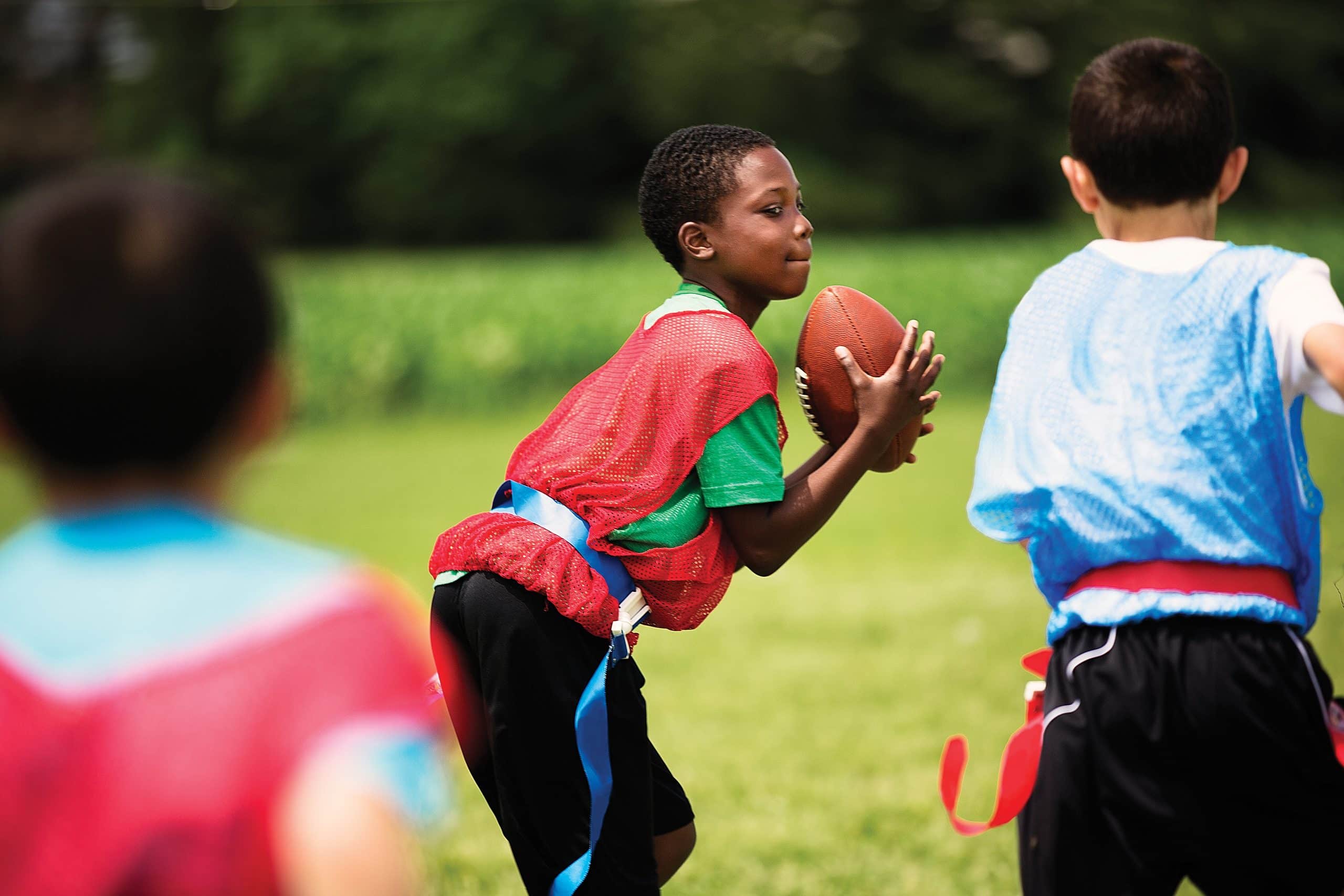 A boy in a red pinnie grips a football on a grass field during a youth flag football game