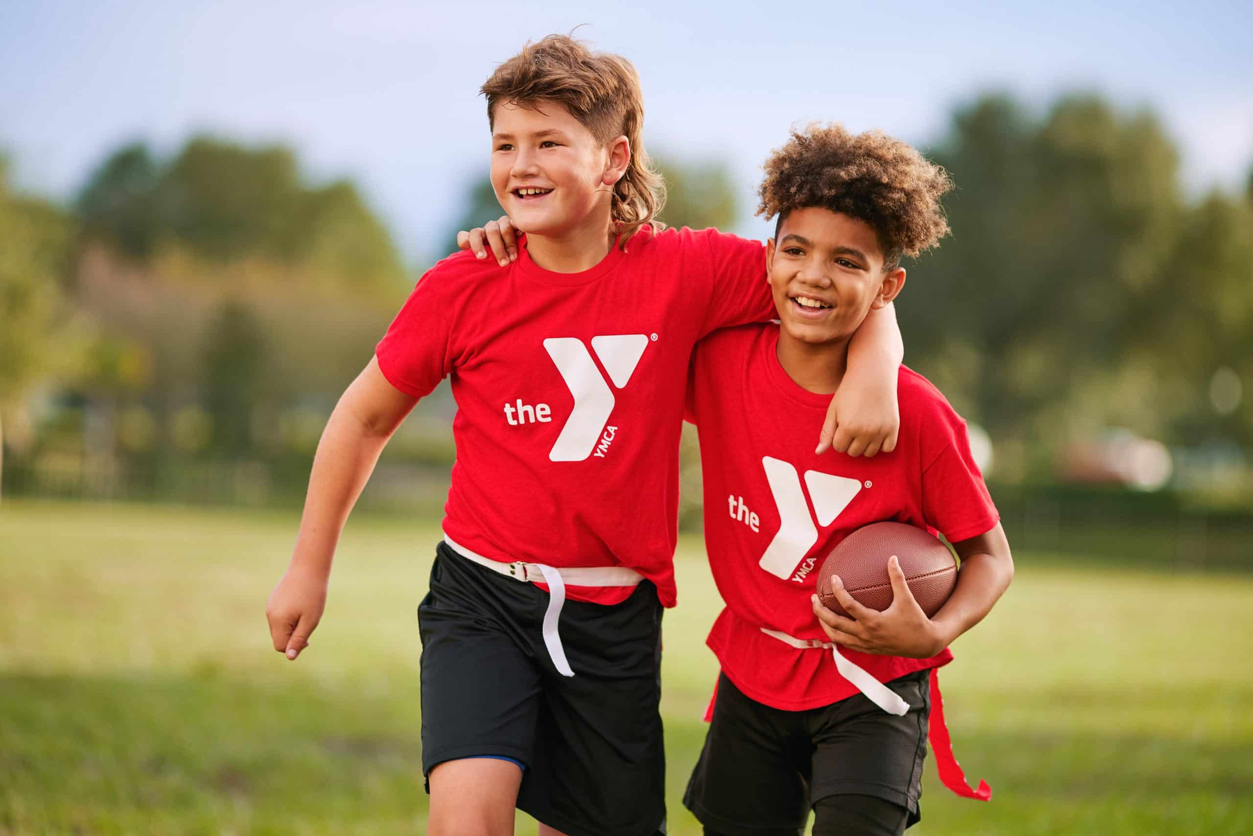 Two boys in red YMCA shirts and flag football belts walk arm in arm across a field, one carrying a football
