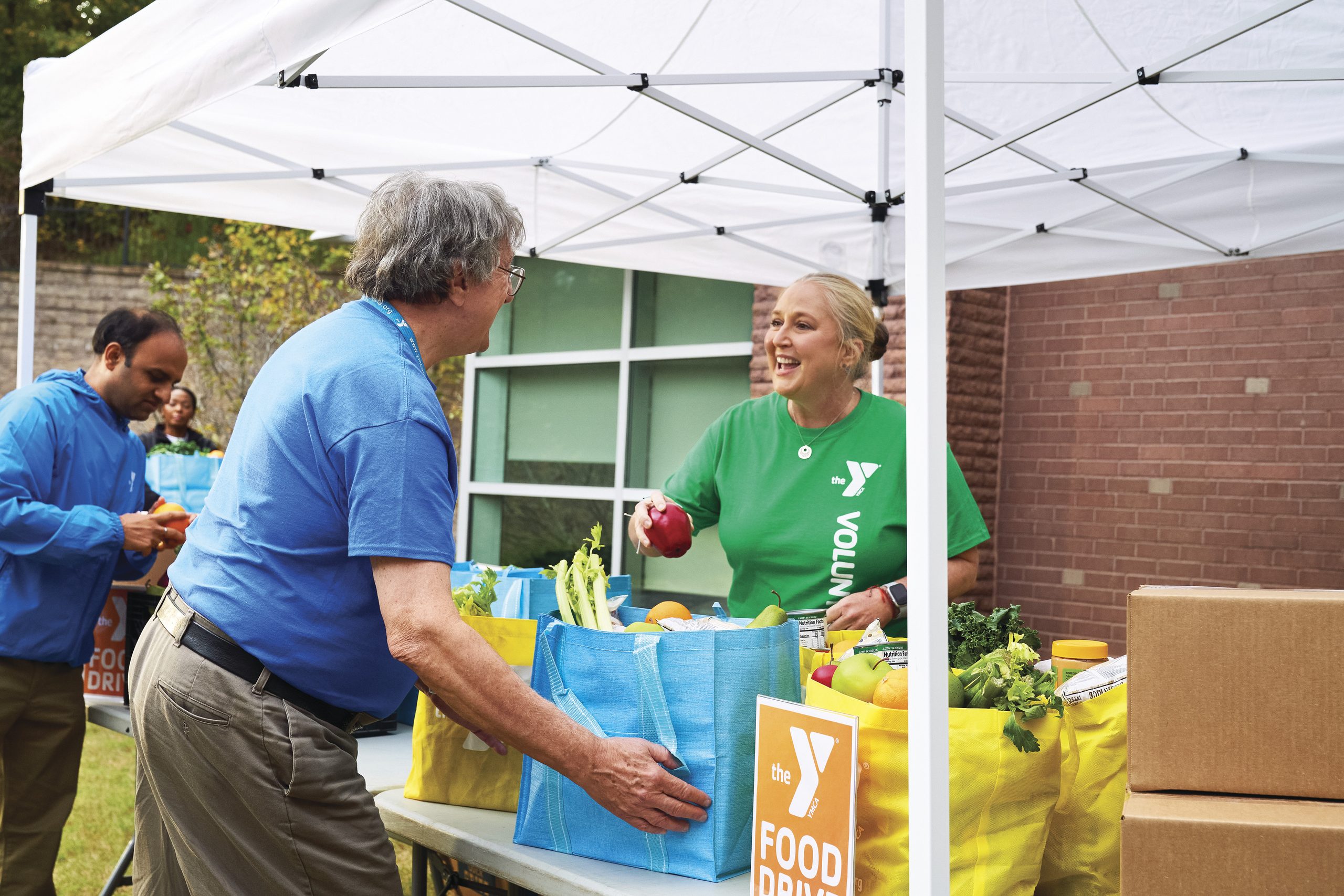YMCA volunteer in a green shirt hands produce to a visitor packing grocery bags at an outdoor food drive