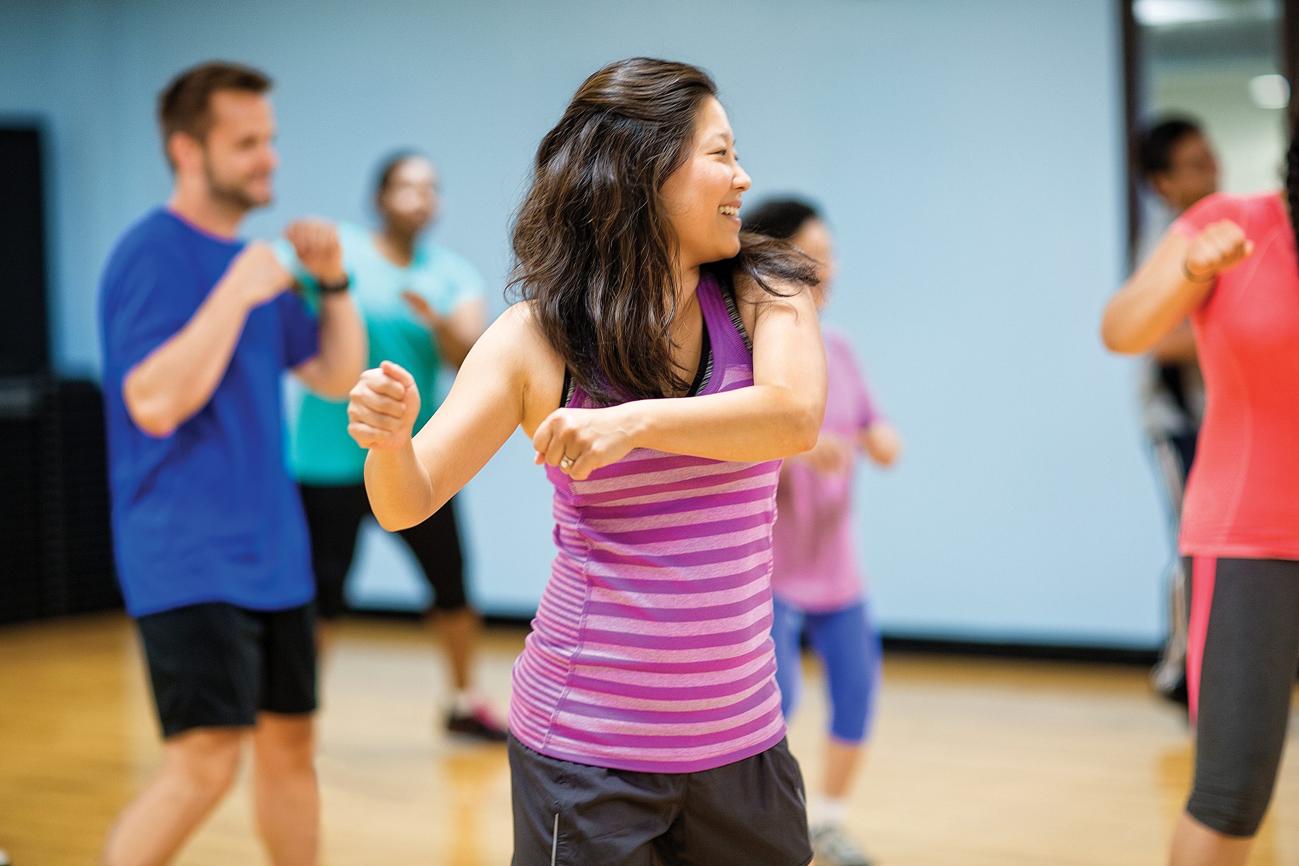 Adults dancing in a group fitness class in a studio, a woman in a purple tank top in front