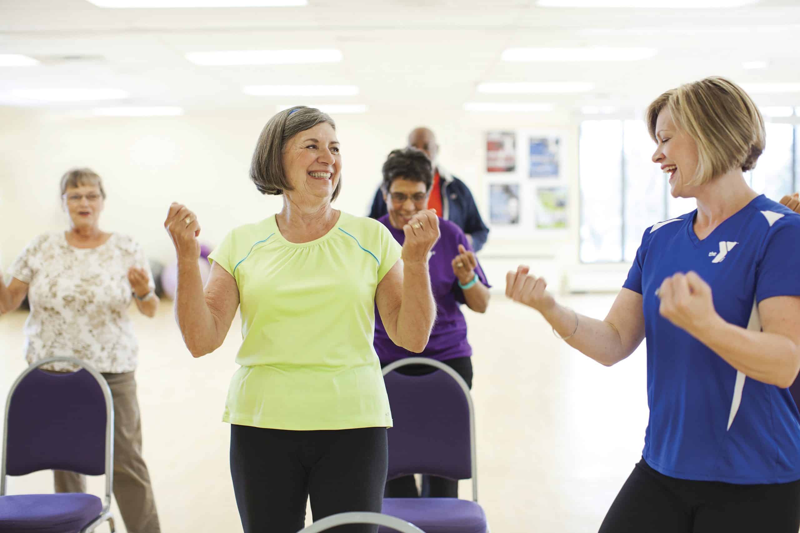 Older adults follow an instructor in a blue YMCA shirt during a standing group fitness class beside chairs