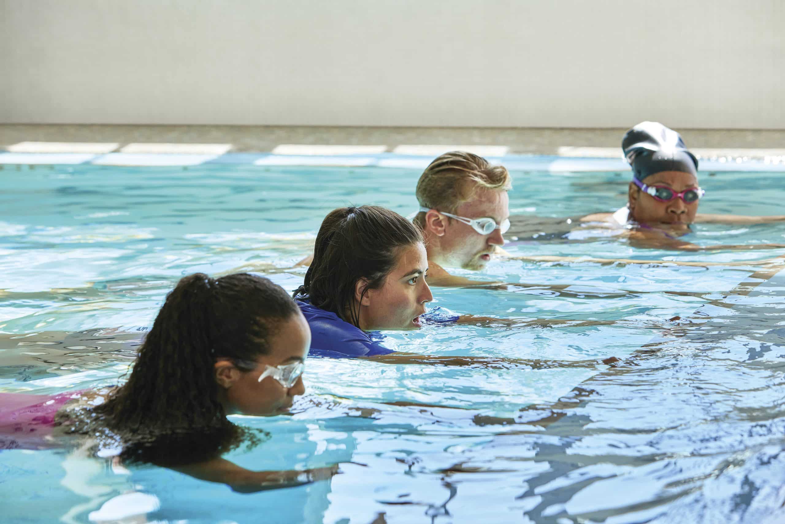 Four adults in goggles line up shoulder to shoulder in an indoor pool during a swim lesson