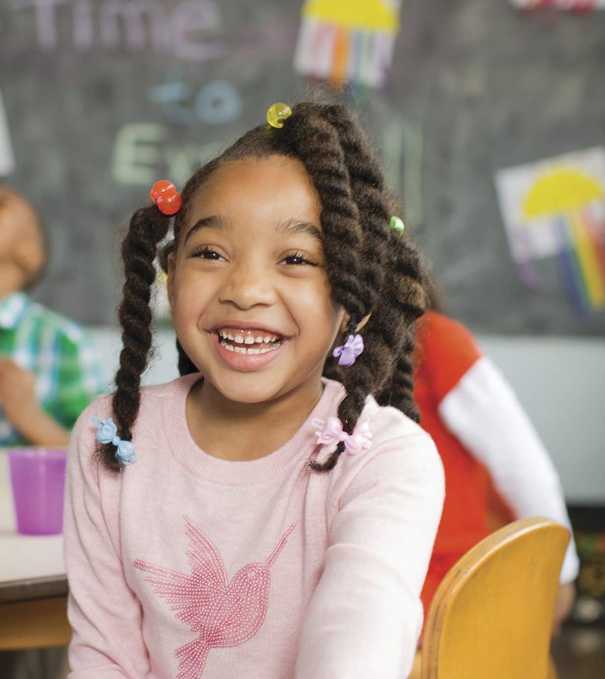A grinning child with beaded braids holds out a purple plate of apple slices at a classroom snack table