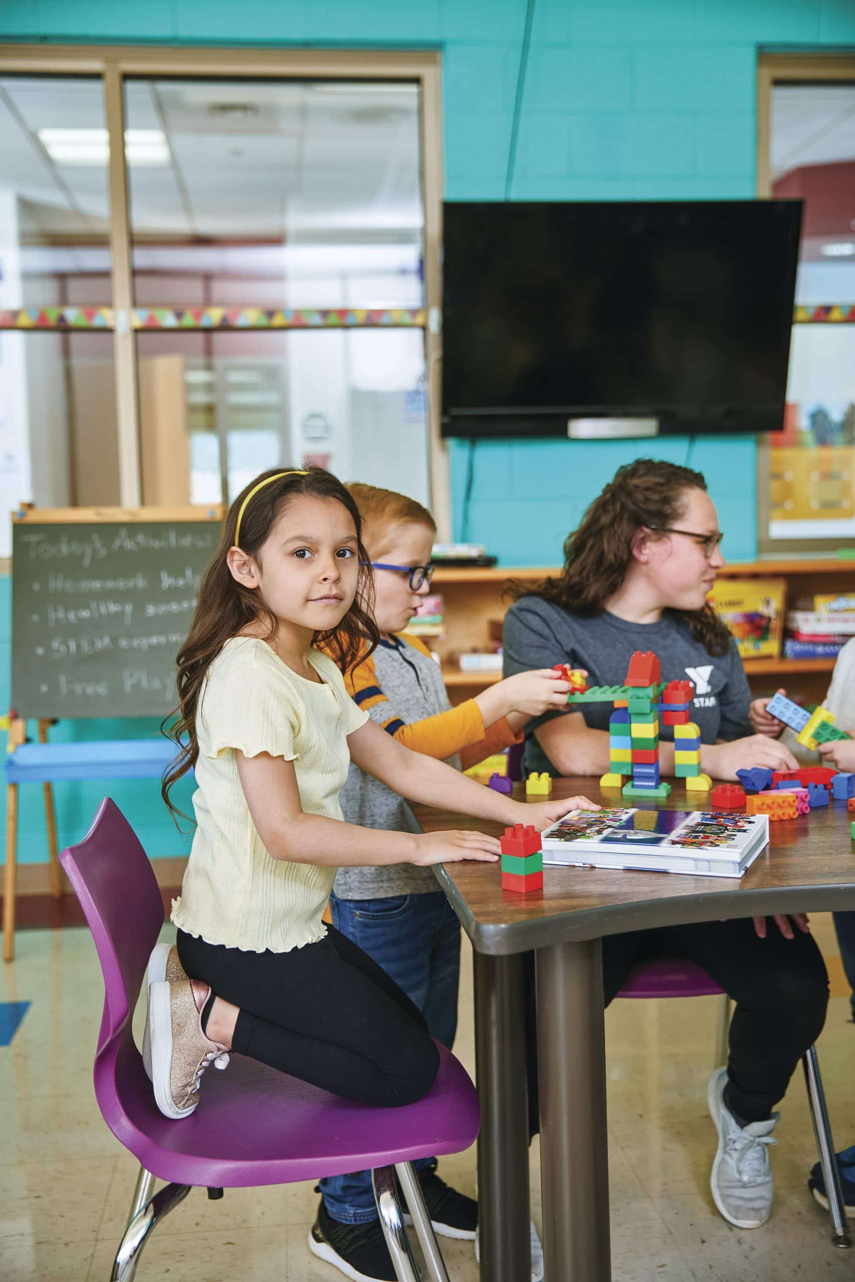 Children build with large plastic blocks at a classroom table while a Y staff member helps