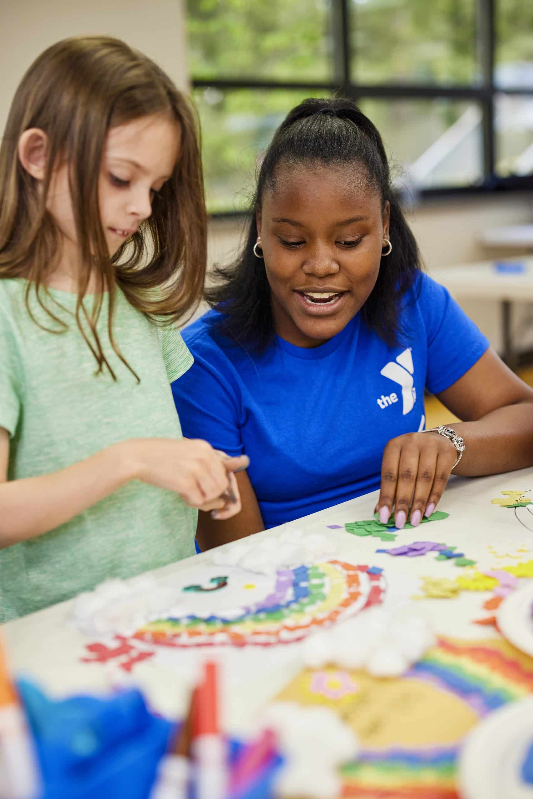 A child and a YMCA staff member in a blue shirt build a rainbow from foam stickers at a craft table