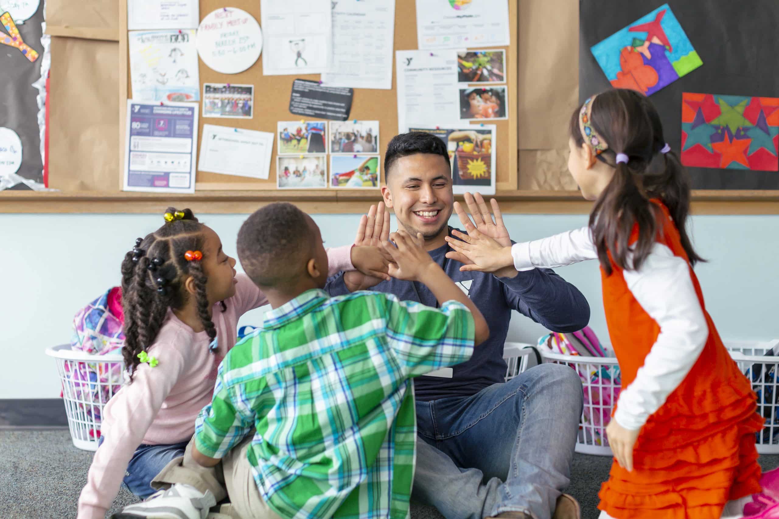 A smiling YMCA staff member gives double high fives to three children in a program room with a bulletin board