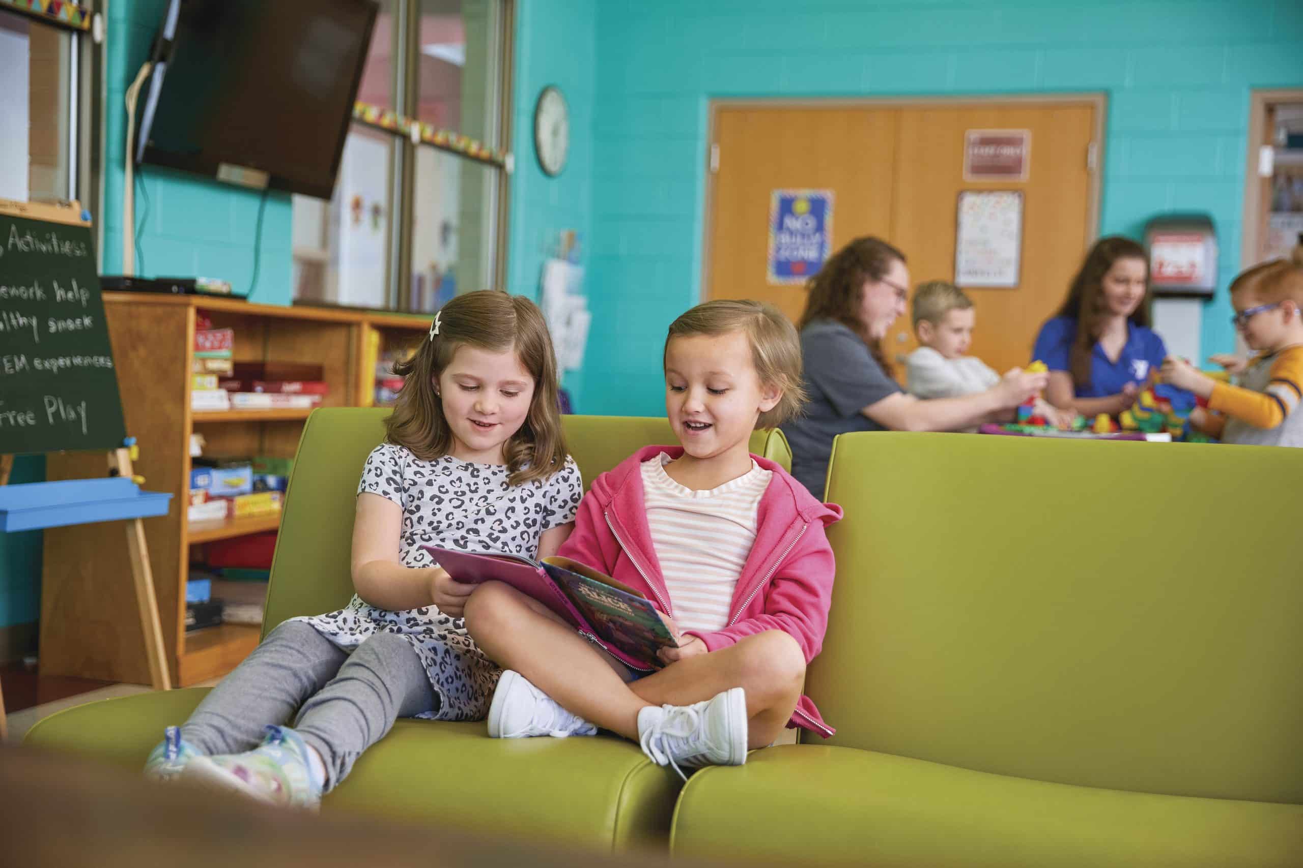 Two young girls sit on a green couch reading a book together in a Y Club classroom