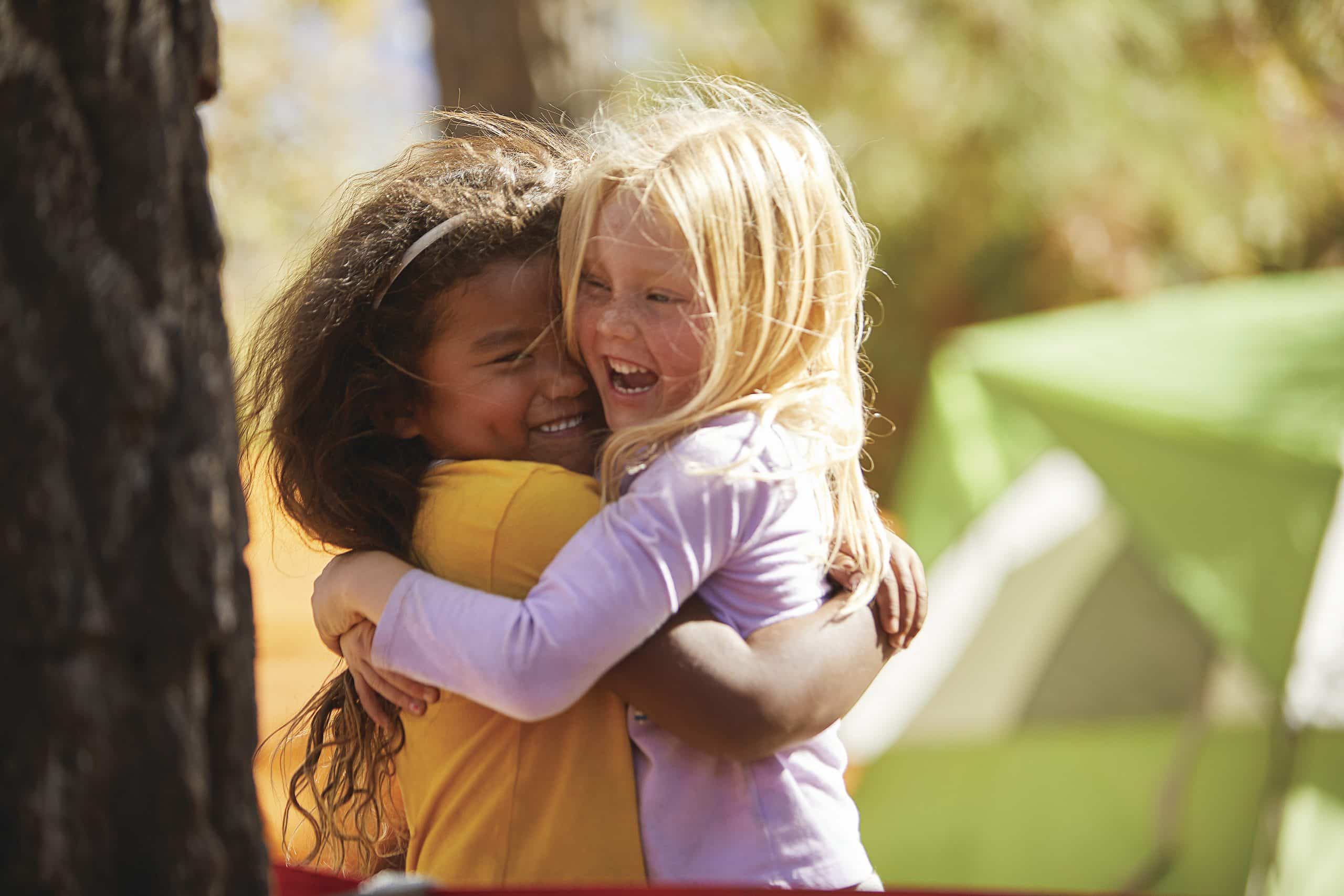 Two young campers hug and laugh outdoors beside a green tent at a wooded campsite