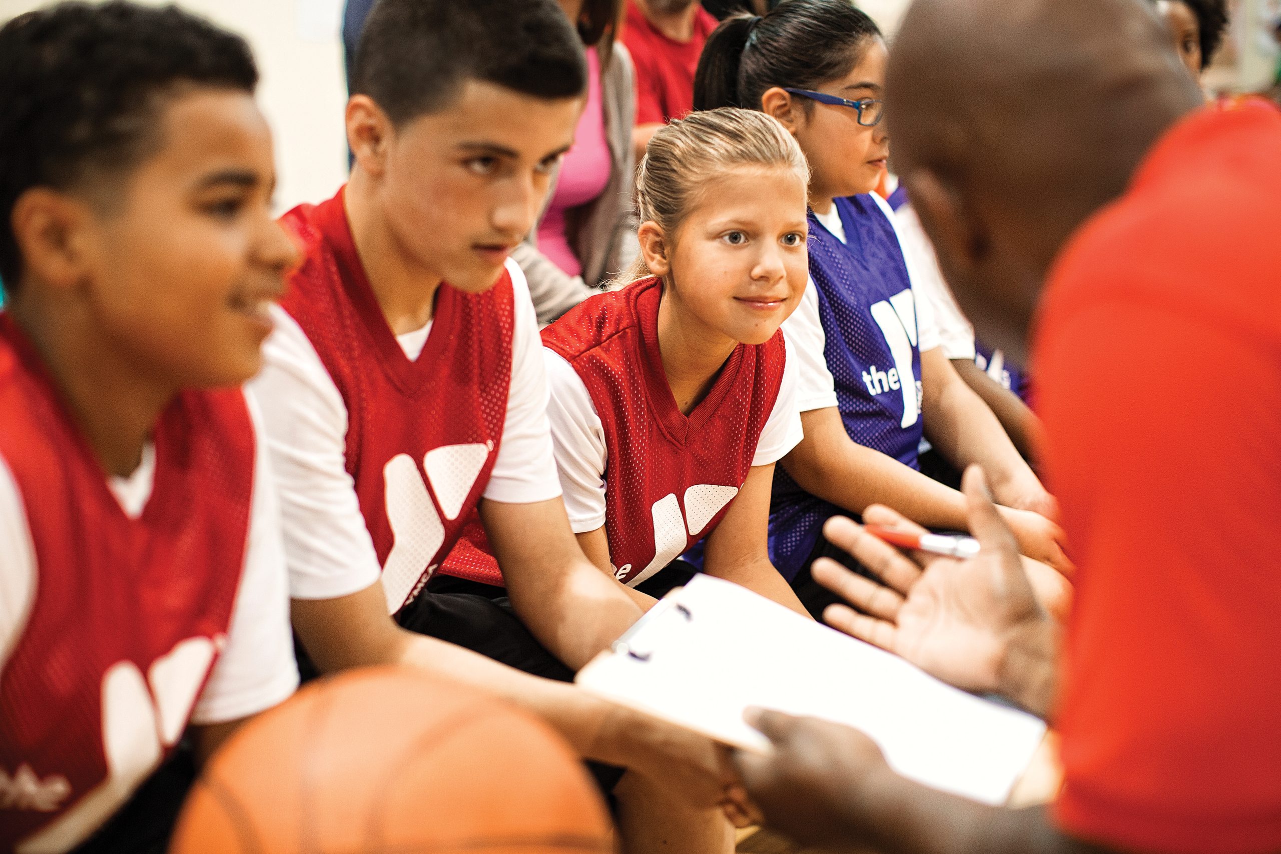 Youth basketball players in red and purple jerseys listen to a coach holding a clipboard