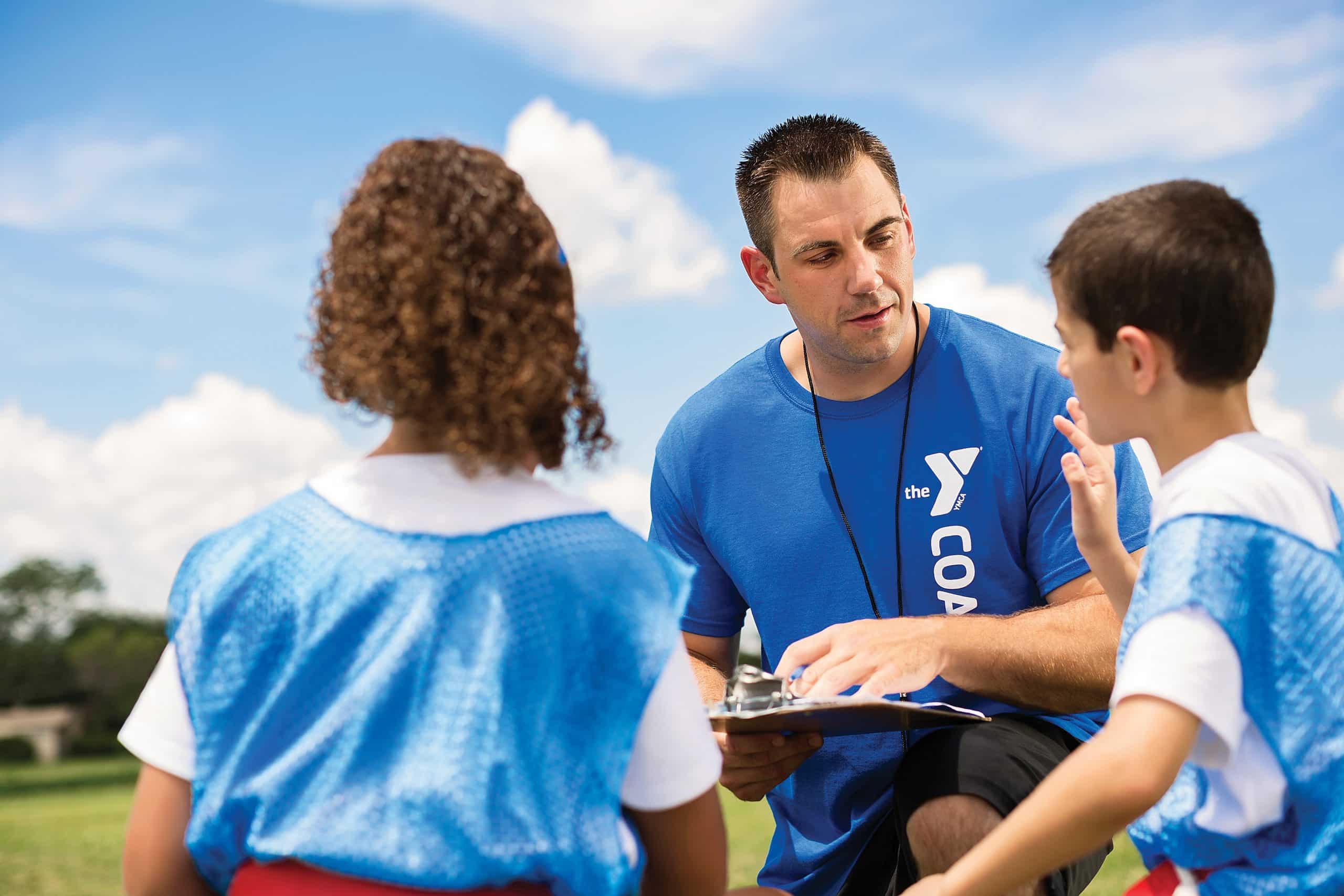 A YMCA coach kneels with a clipboard to talk through a play with two young players in blue pinnies