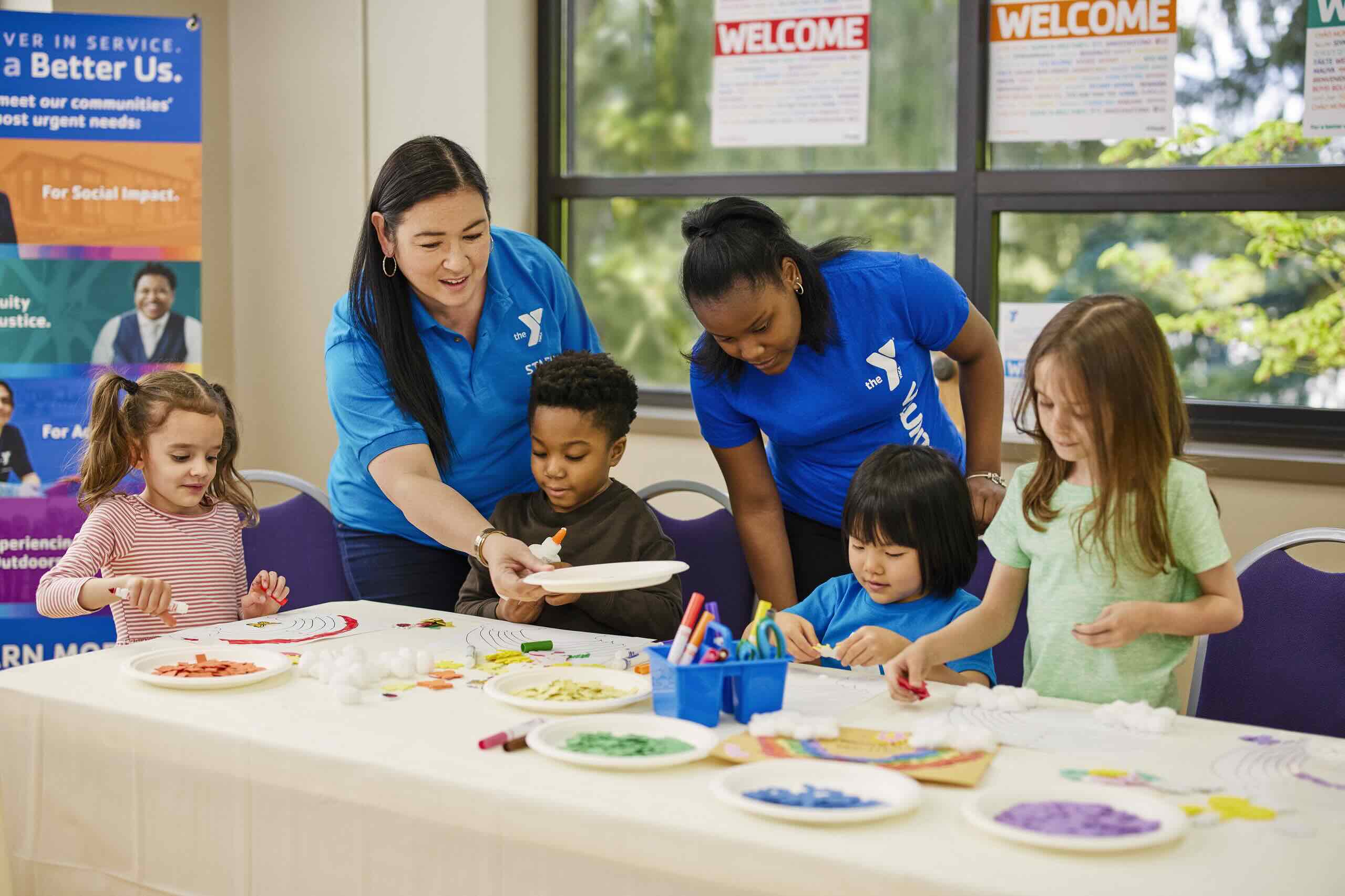 Four young children work on art and crafts at a table with the assistance of two Y staff members.