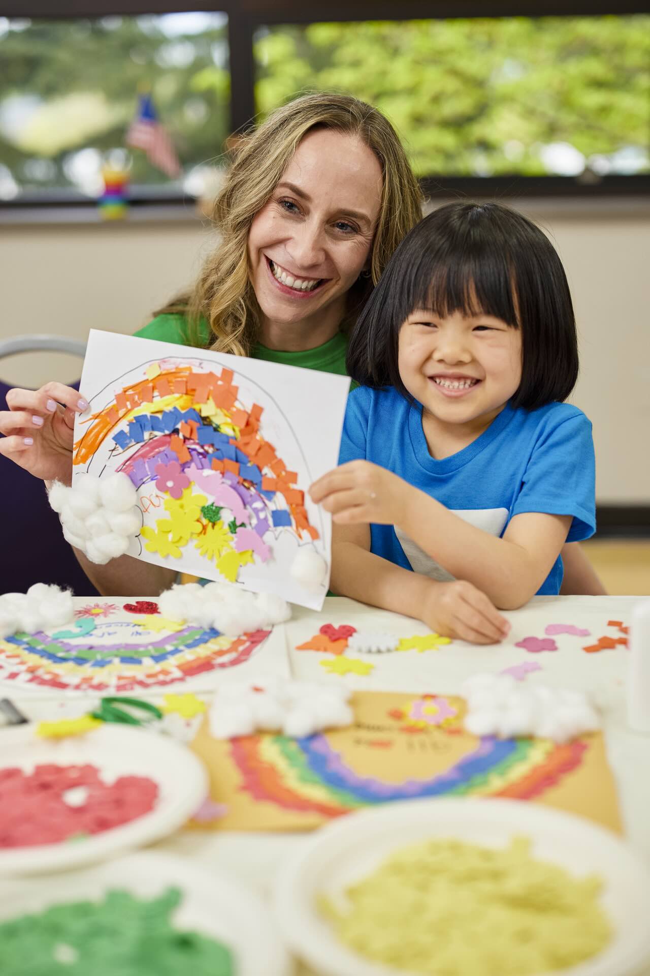 A young girl smiles while holding up a rainbow piece of art that a Y staff member helped her create.