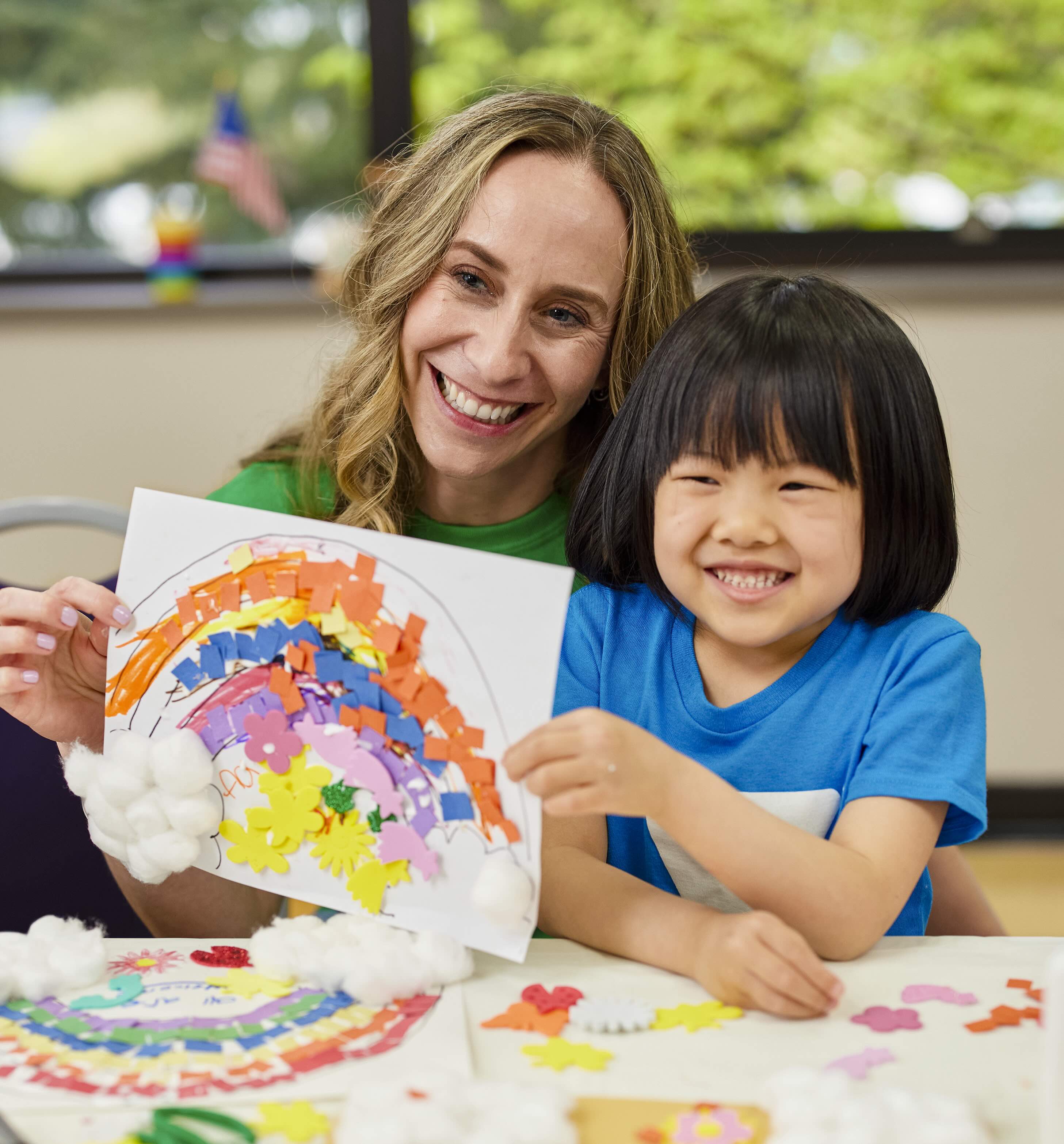 Teacher and preschool student smiling and holding rainbow artwork.