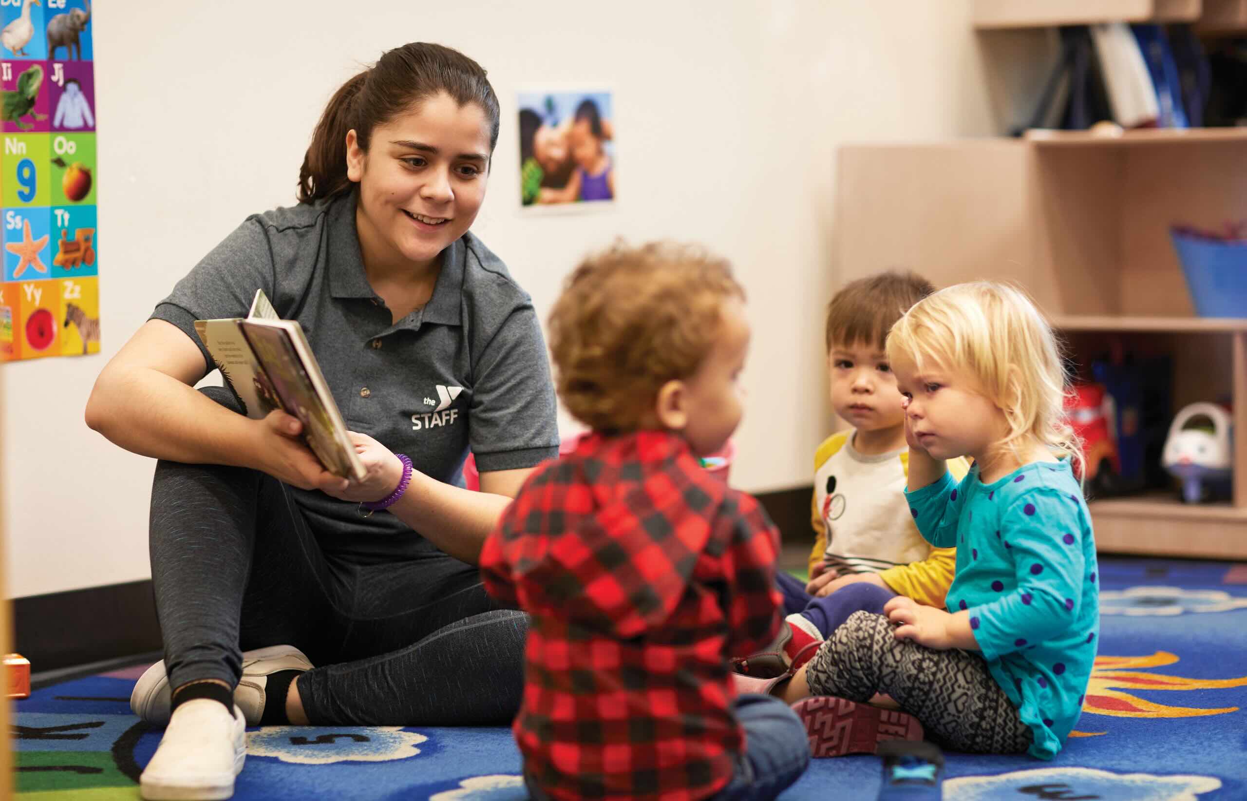 A Y staff member sits on the floor with three young children while reading a story book to them.