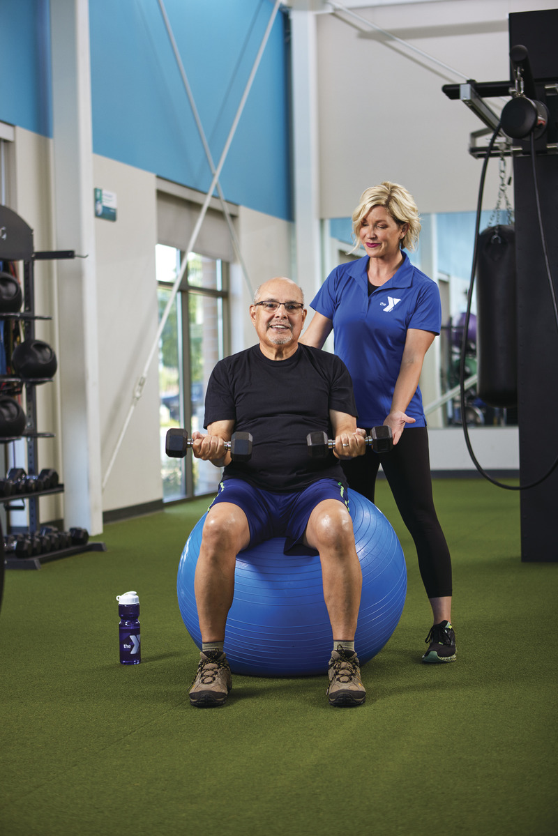 Older gentleman doing bicep curls on a medicine ball with guidance from Kansas City YMCA personal trainer