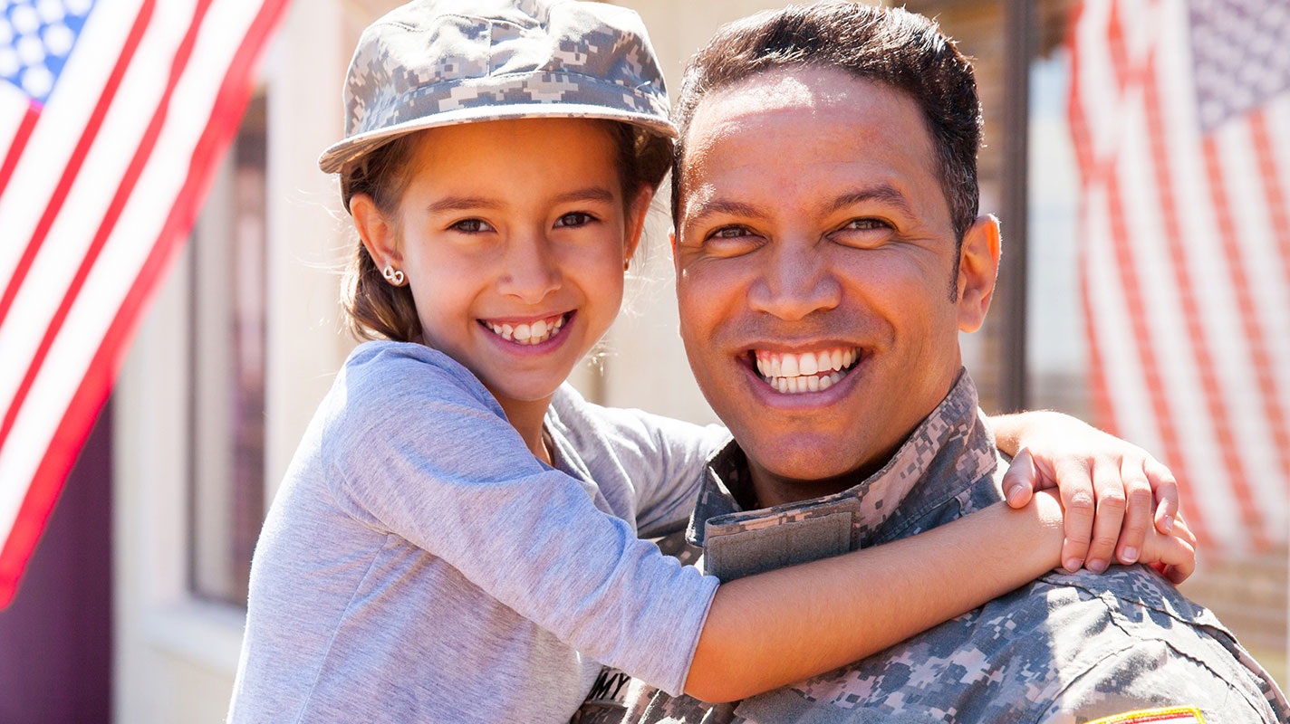 Dad in military uniform holding daughter, both smiling, with an American flag in the background