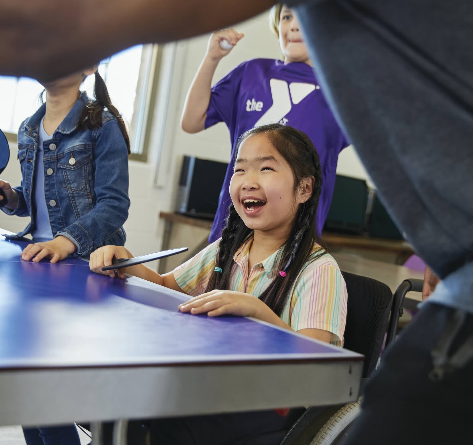 Smiling girl in wheelchair holding ping pong paddle playing with other students.