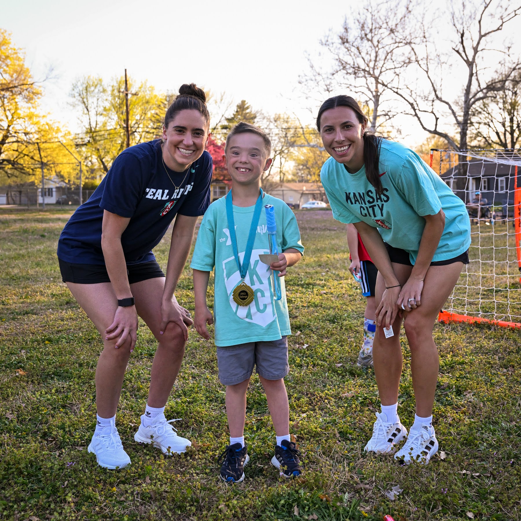 Two KC Current soccer players stand with Challenger soccer participant in the middle, who is wearing a medal and YMCA KC Current soccer shirt
