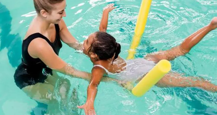 Young girl floating on a pool noodle while swimming toward a swim instructor. 