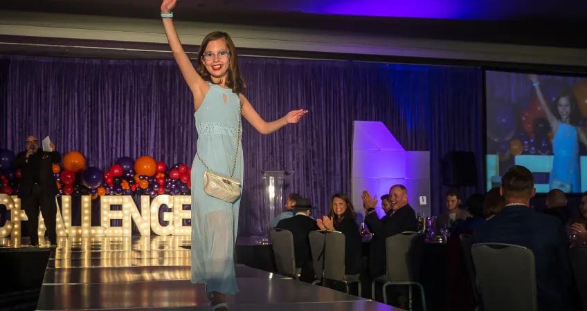 Challenger athlete Maddie Richardson struts down the runway in a floor length blue gown and a cute white purse. The audience is smiling and clapping.