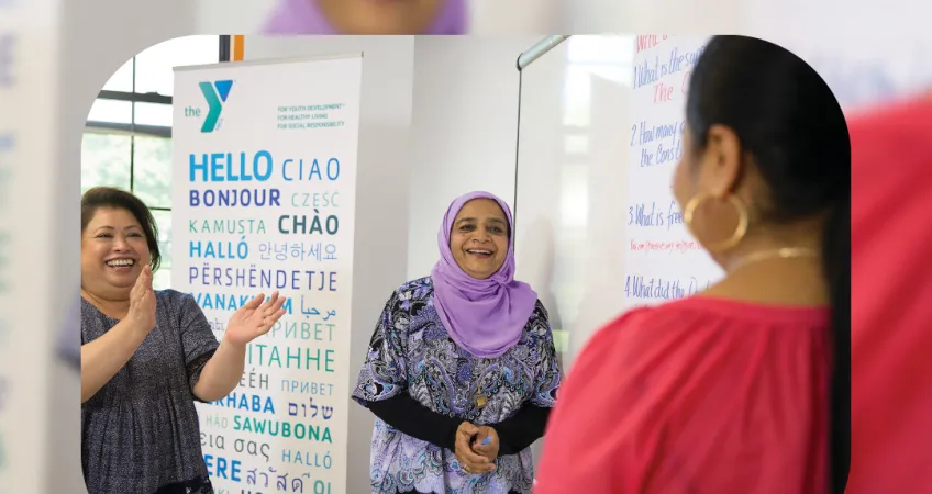 Kansas City YMCA graphic featuring two women smiling and clapping their hands to a person whose back is facing the camera. They are in front of a YMCA sign saying "hello" in many different languages. There's a bottom text banner saying "Immigrant Heritage Month".